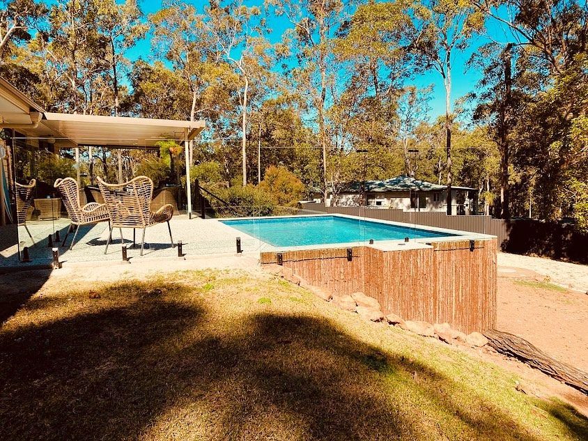 Swimming pool with wooden border and patio furniture, surrounded by trees. — HinterCoast Pools In Tin Can Bay, QLD