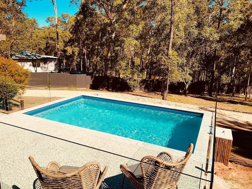 Rectangular pool with blue water, surrounded by concrete patio and two wicker chairs; trees in background. — HinterCoast Pools In Wolvi, QLD