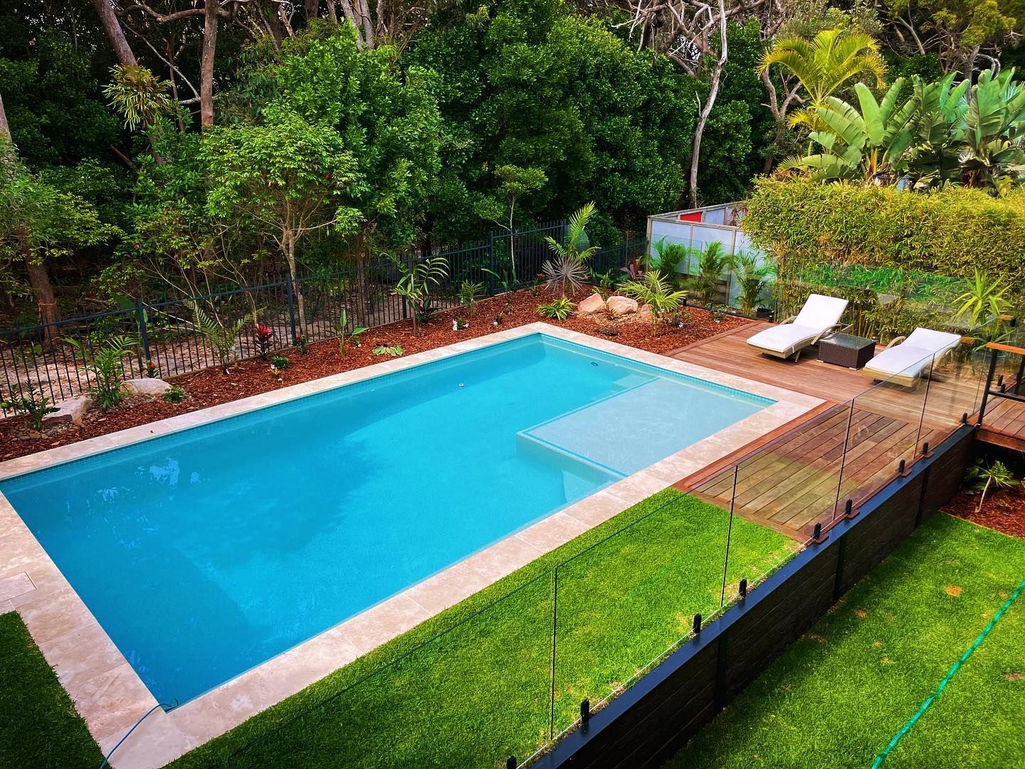 Rectangular pool with turquoise water, surrounded by grass and wooden deck with two lounge chairs, lush greenery in the background. — HinterCoast Pools In Wolvi, QLD