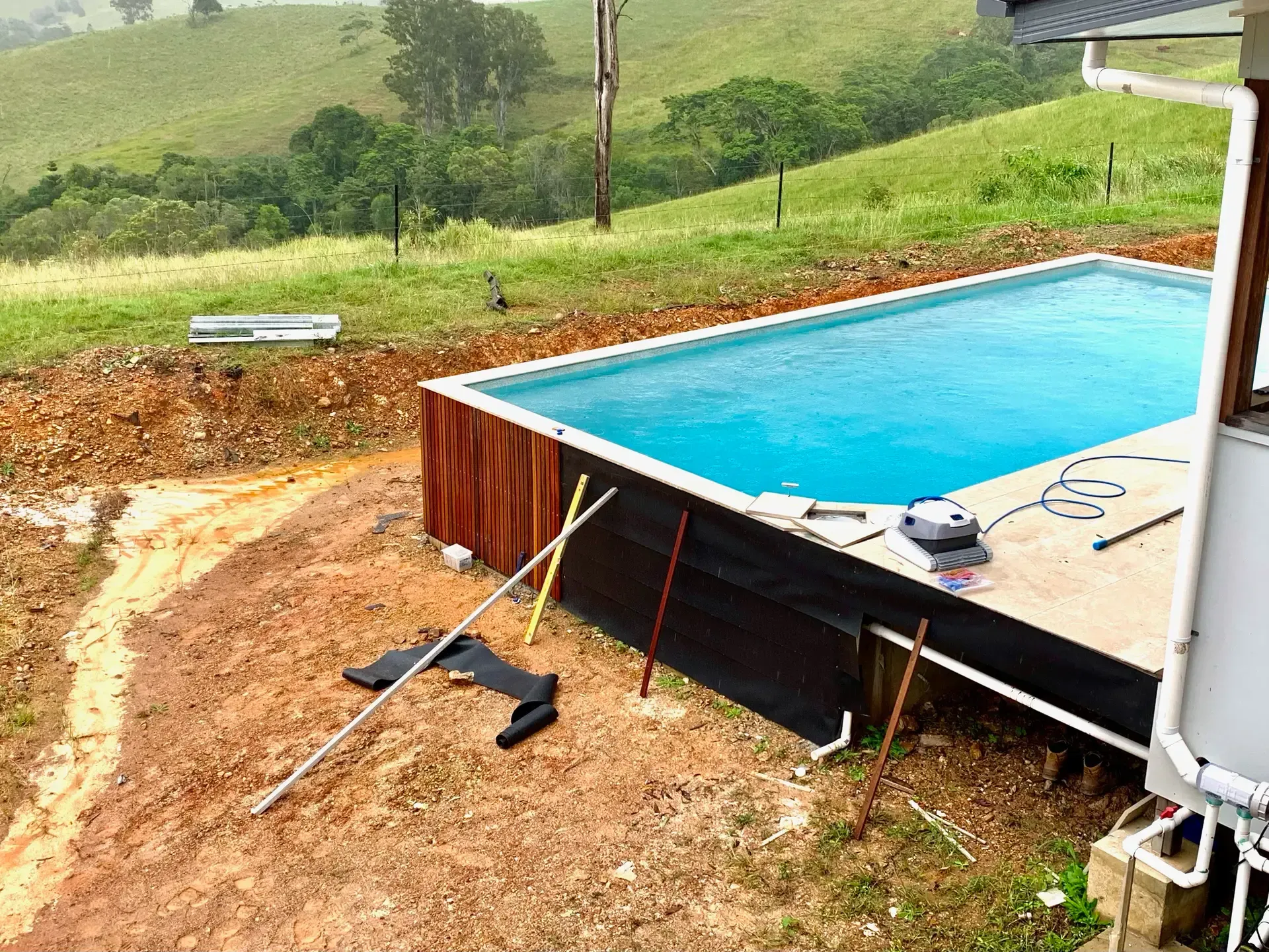 A partially built rectangular pool with blue water next to a building on a grassy hill. Construction equipment visible. — HinterCoast Pools In Wolvi, QLD
