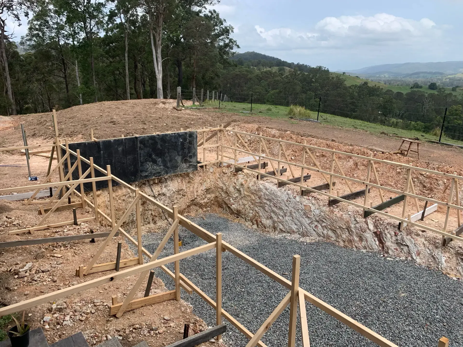 Construction site on a hillside with wooden frameworks, gravel, and a dark wall. Trees and a distant valley are visible. — HinterCoast Pools In Curra, QLD