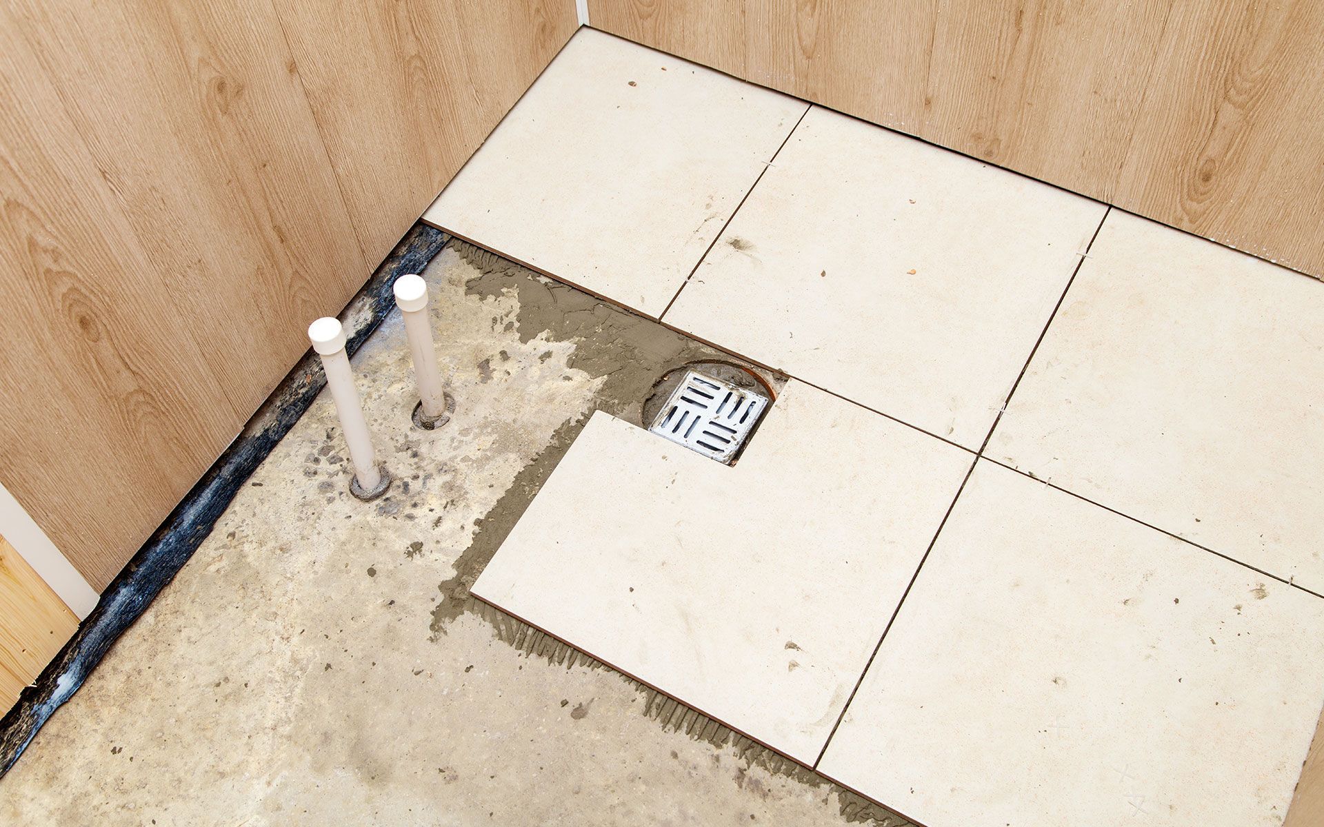 Bathroom floor tiling in progress: Beige tiles laid around a drain and pipes, showing the substrate.