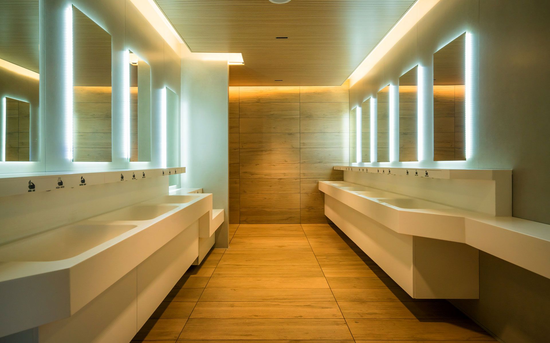 Modern public restroom with several white sinks, mirrors with bright lights, and wooden floors.