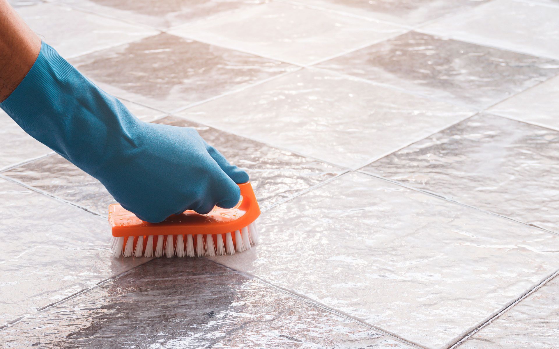 Hand in blue glove scrubbing checkered tile floor with orange brush.