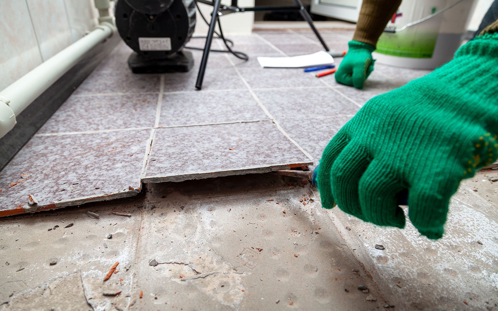 Hands in green gloves lifting damaged floor tile.