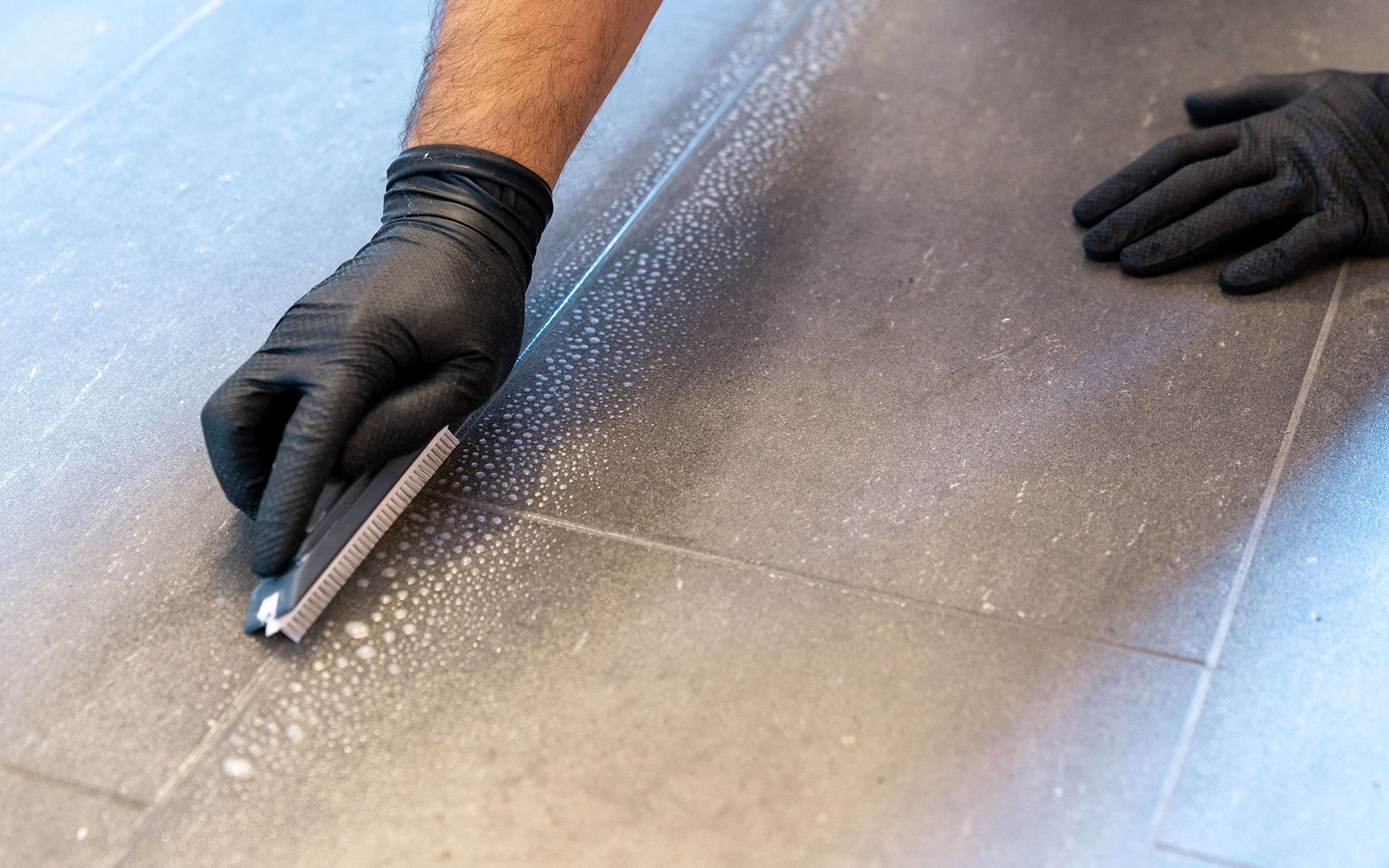 Person wearing black gloves cleaning tiled floor with a scraper.