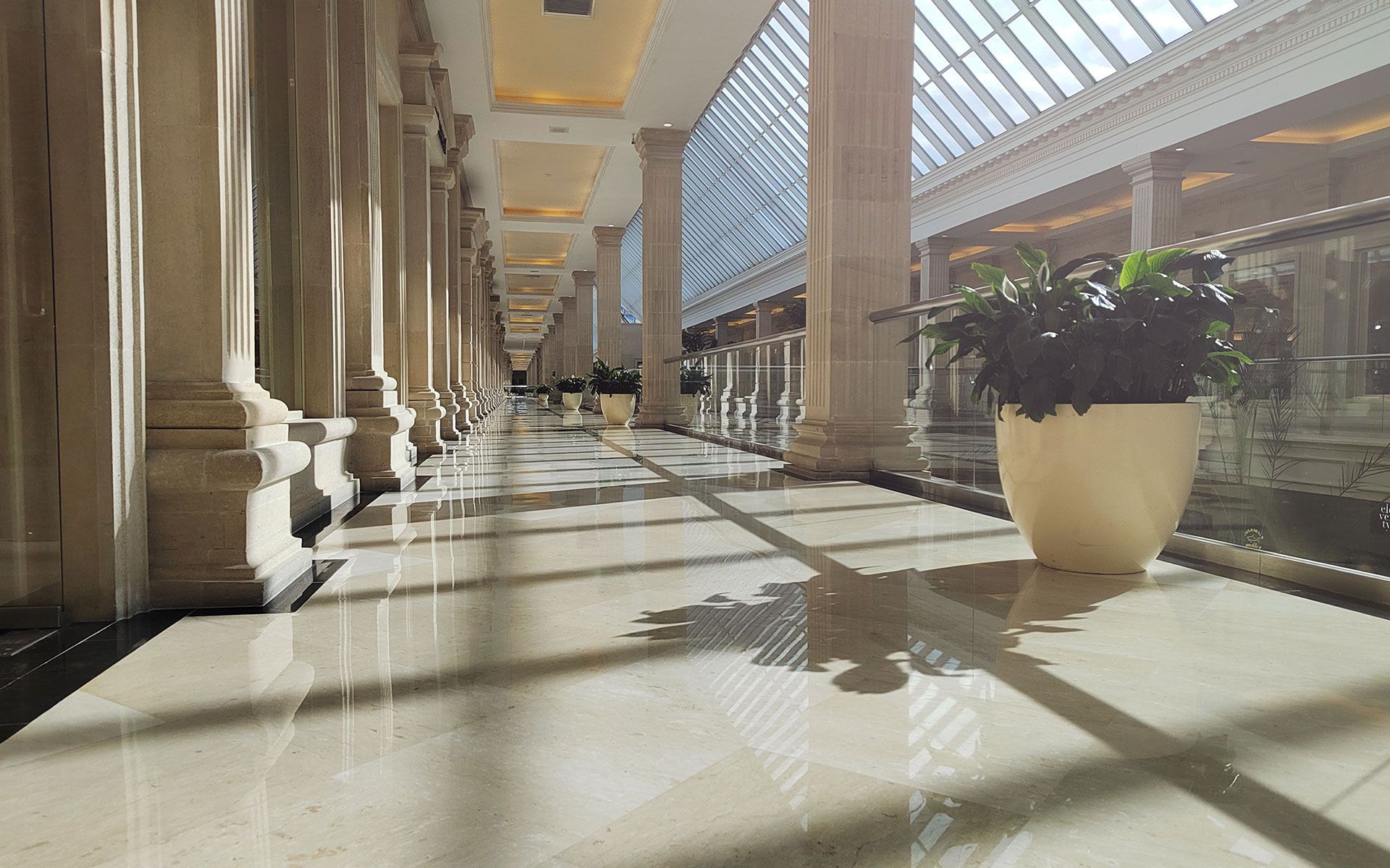 Long hallway with marble floor, sunlight, pillars, and potted plants.