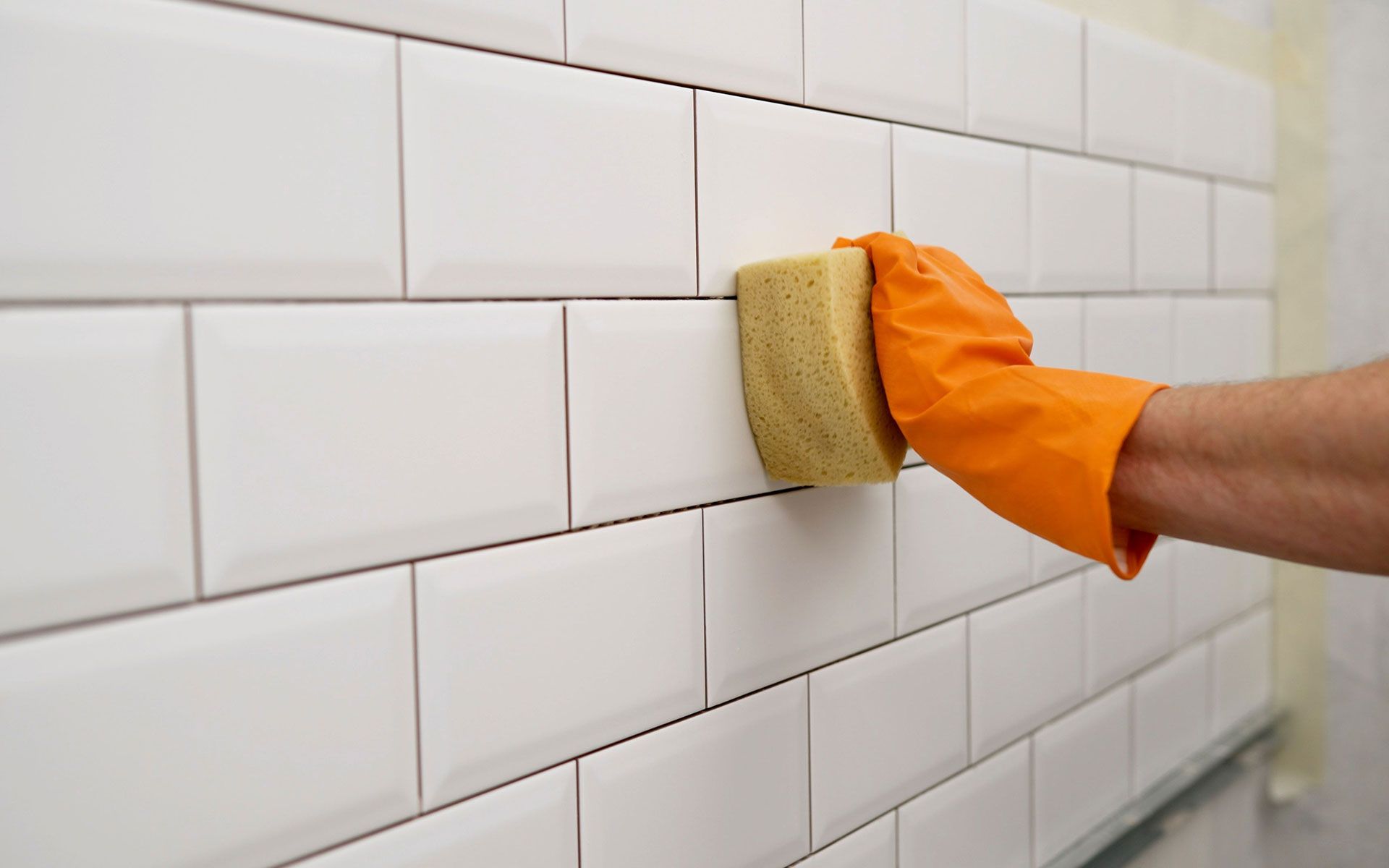 Hand in orange glove wiping white tile wall with a yellow sponge.
