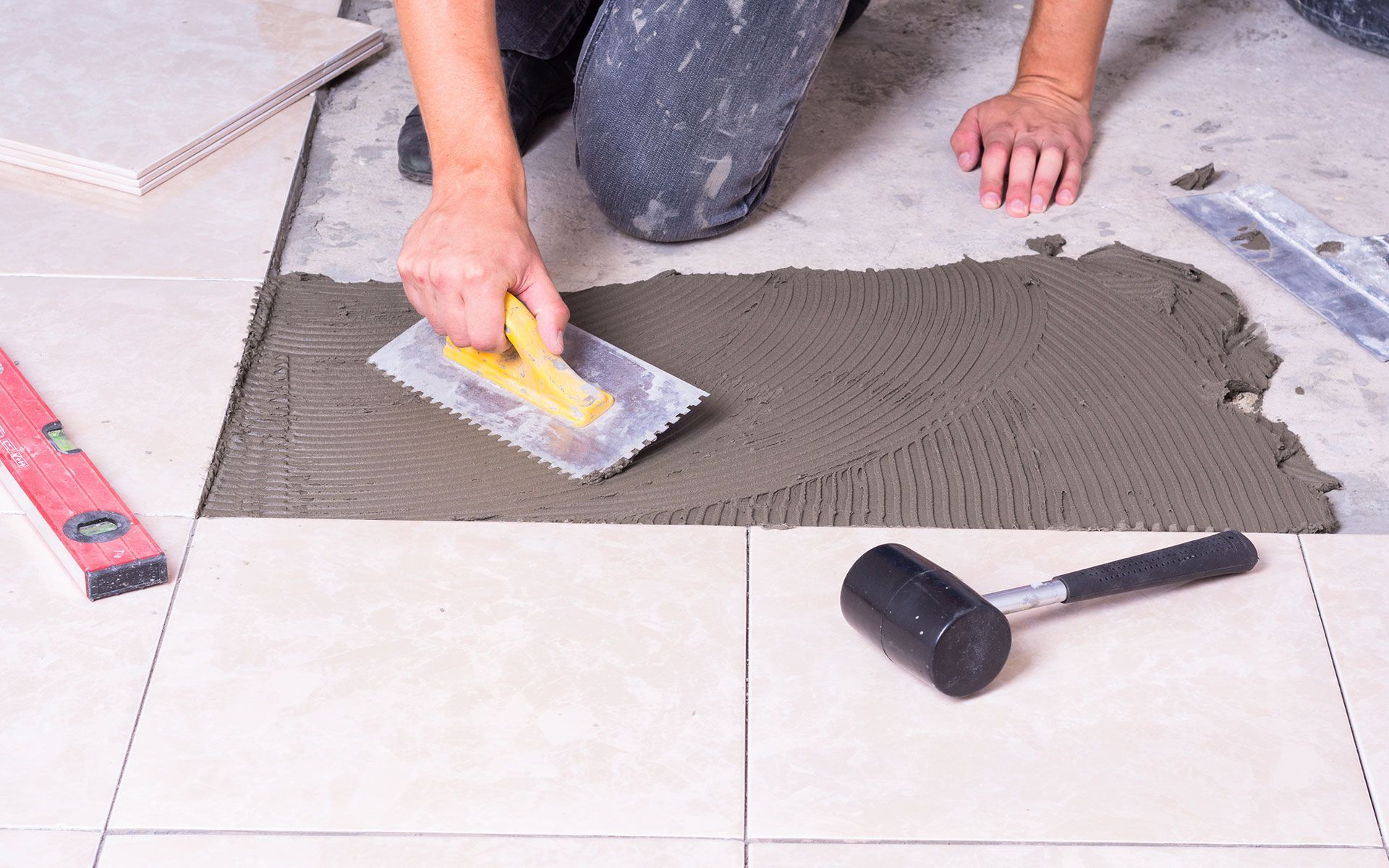 Person tiling a floor, using a notched trowel to spread mortar, near a level and rubber mallet.