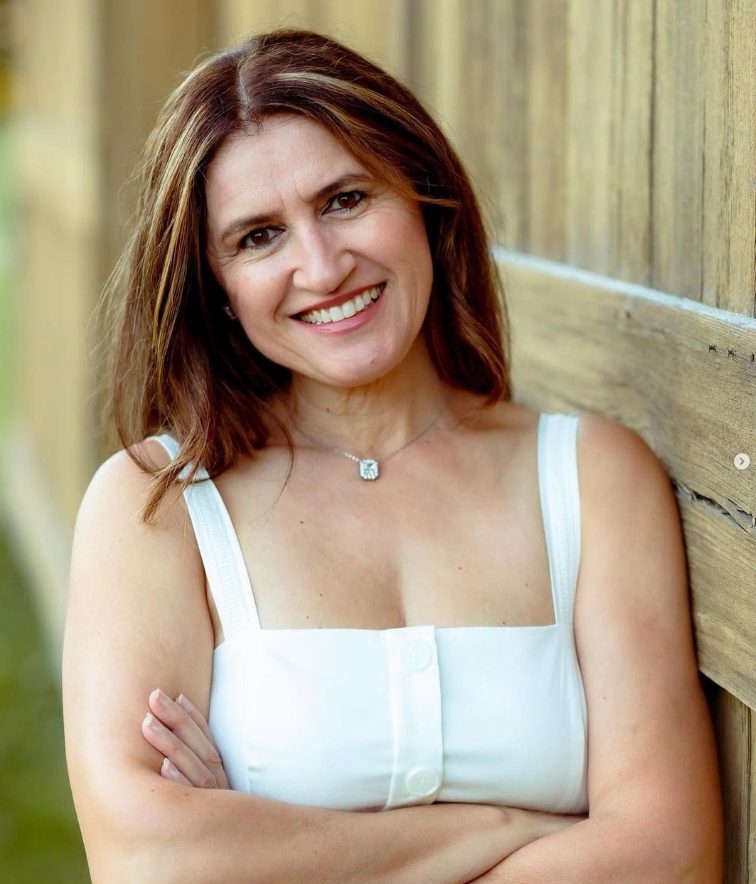 Fifi Papdopoulos in a white tank top is leaning against a wooden wall with her arms crossed and smiling. Fifi Papdopoulos in a white tank top is leaning against a wooden wall with her arms crossed and smiling.