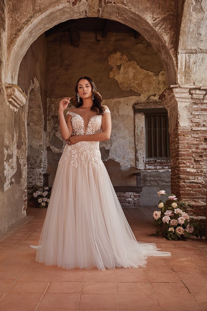 Woman in a wedding dress poses inside an arched stone building; she wears a tulle skirt and embroidered bodice.