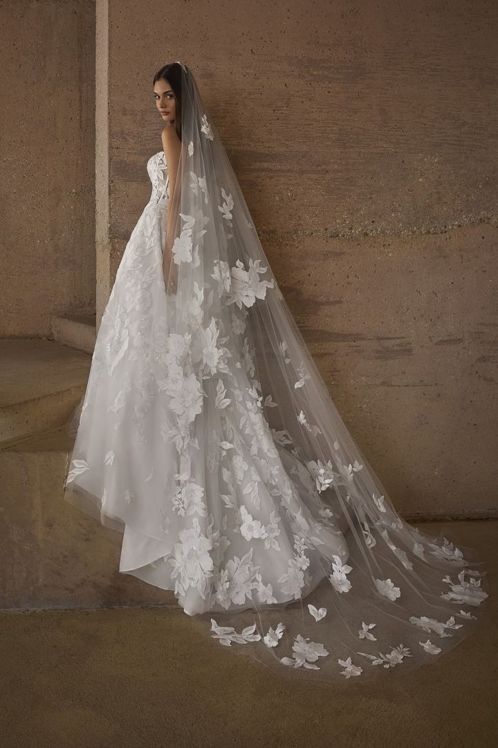 Bride in a white floral lace wedding gown and veil, standing near a stone wall, looking over her shoulder.