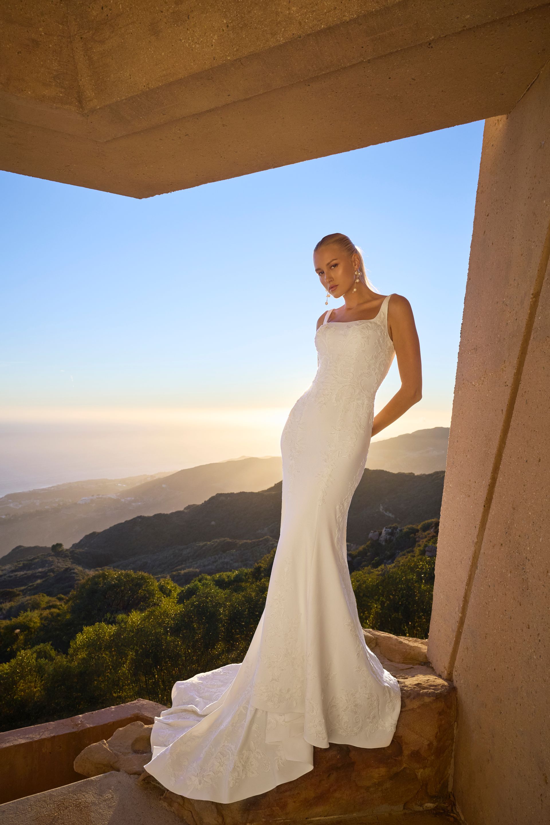 A woman in a white wedding dress is standing in front of a window overlooking a mountain range.