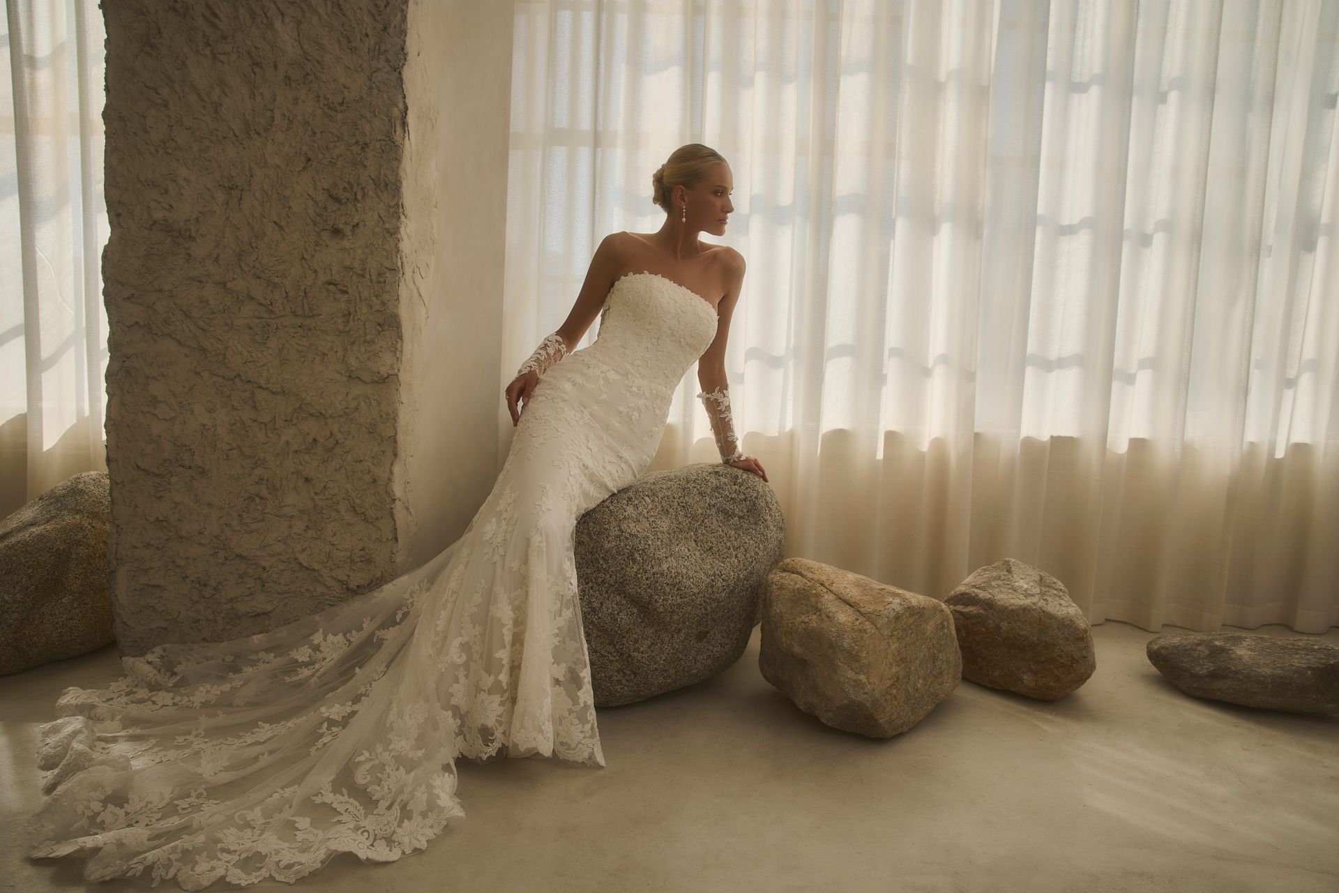 Bridesmaids, in white dresses, sit on a ledge with the bride, wearing her train, adjusting hair; outside, stone building.