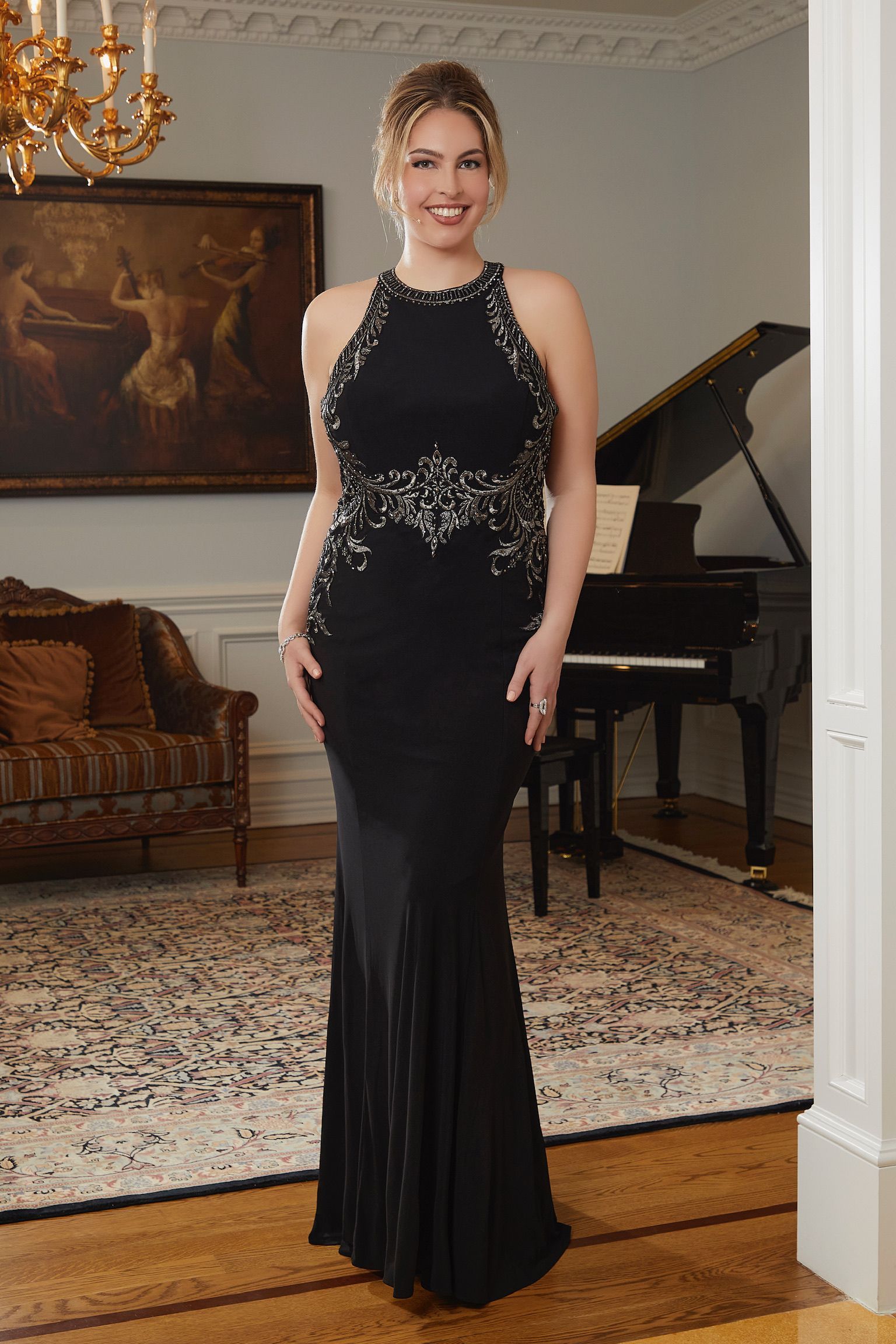 A woman in a black dress is standing in a living room next to a piano.