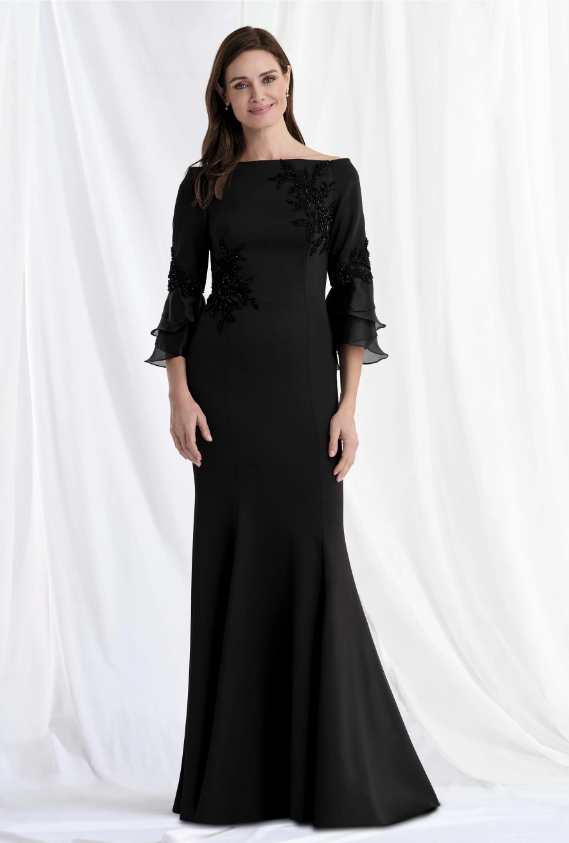 Woman in a long black formal gown with floral accents and fluted sleeves, standing in front of white draped fabric.