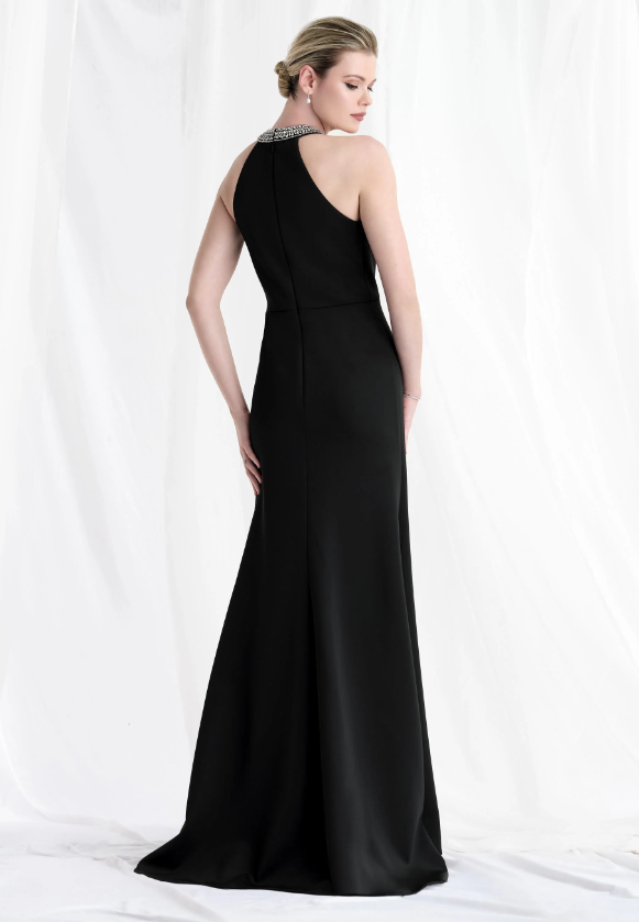 Woman in black evening gown, back view, standing against a white draped background. Dress has embellished neckline and a slight train.