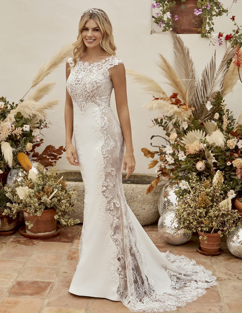 Woman in white lace wedding dress, smiling. Outdoor setting, floral backdrop.