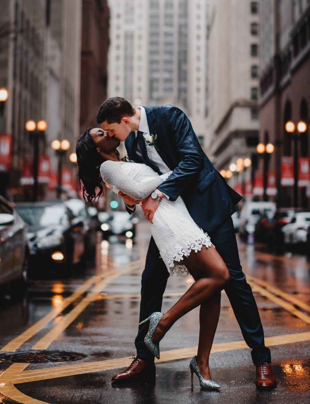 Newlyweds embrace on a city street, rain-slicked pavement. She wears a white dress, he a blue suit.