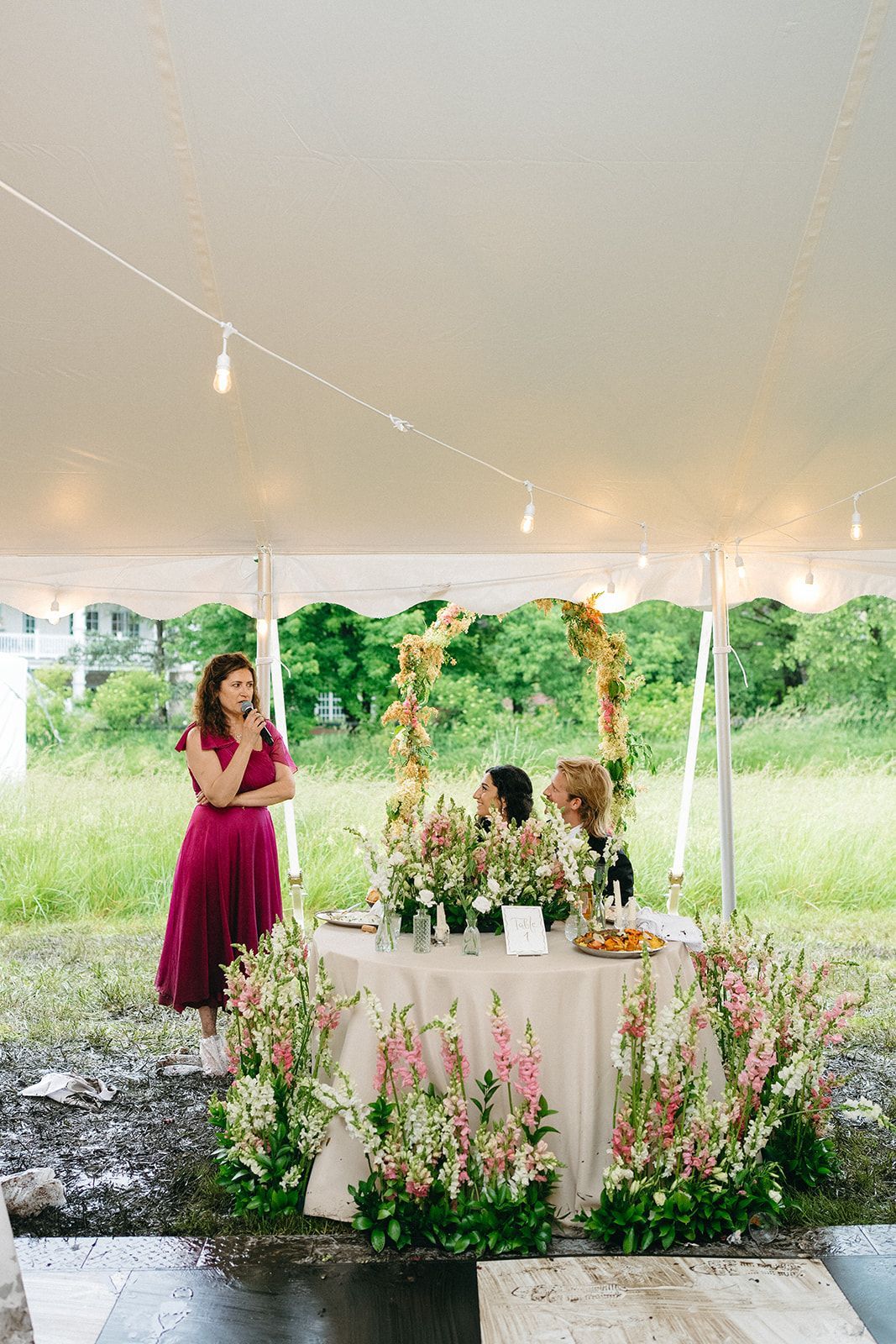 A woman is standing in front of a table with flowers on it under a tent.