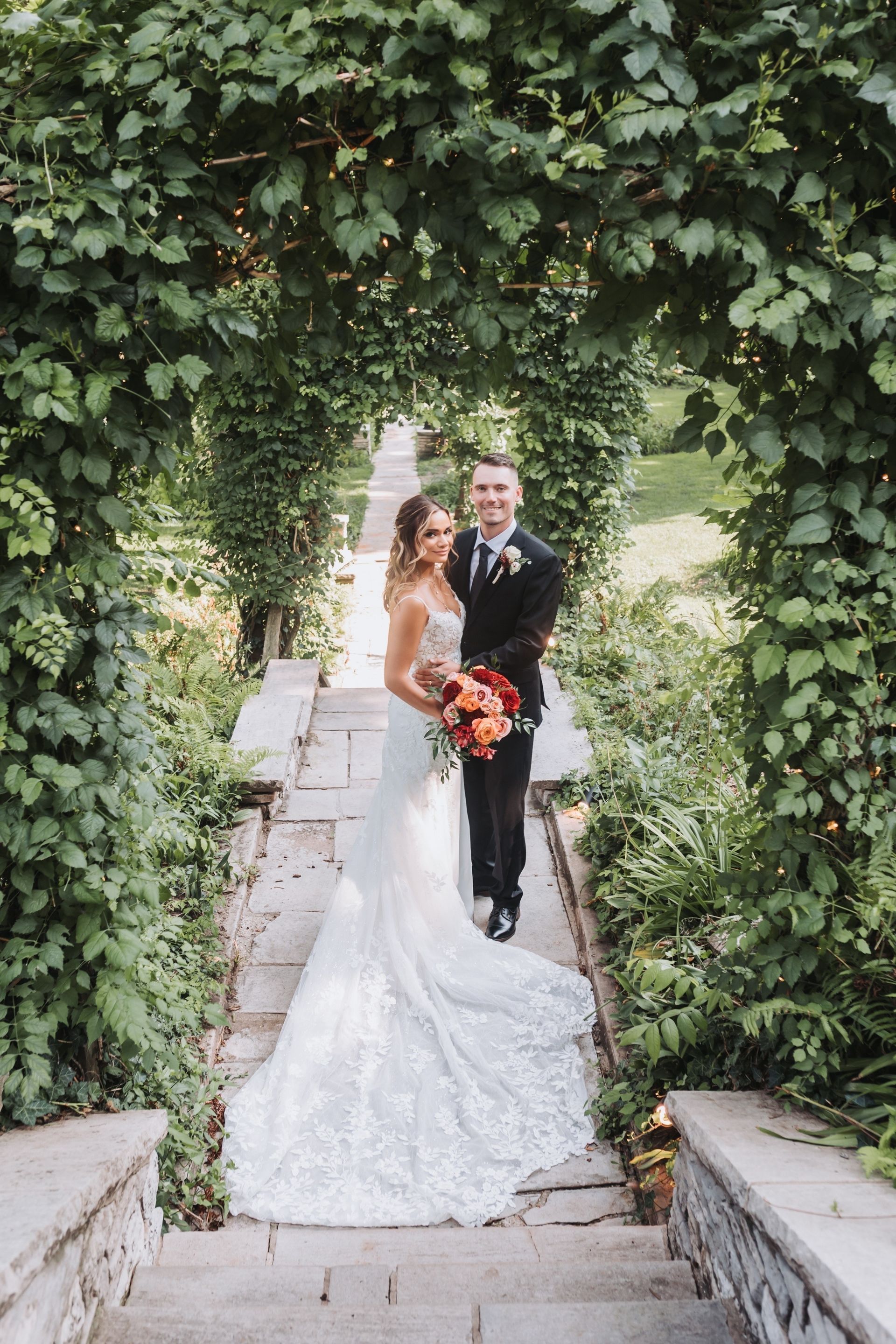 Bride and groom under vine-covered archway on stone steps, holding flowers, smiling, sunny garden setting.