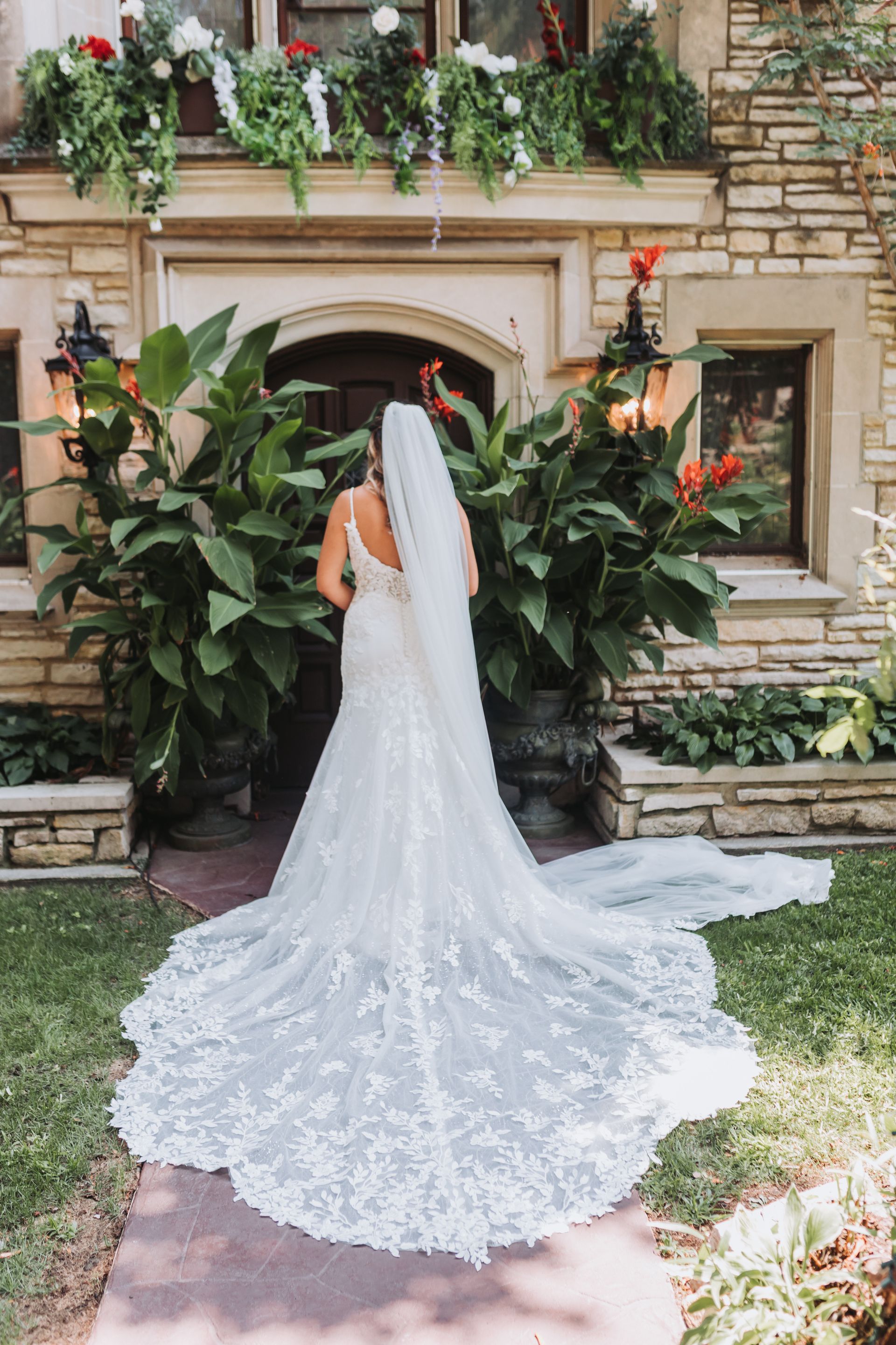 Bride in a lace wedding dress, standing in front of a stone building with floral decorations.