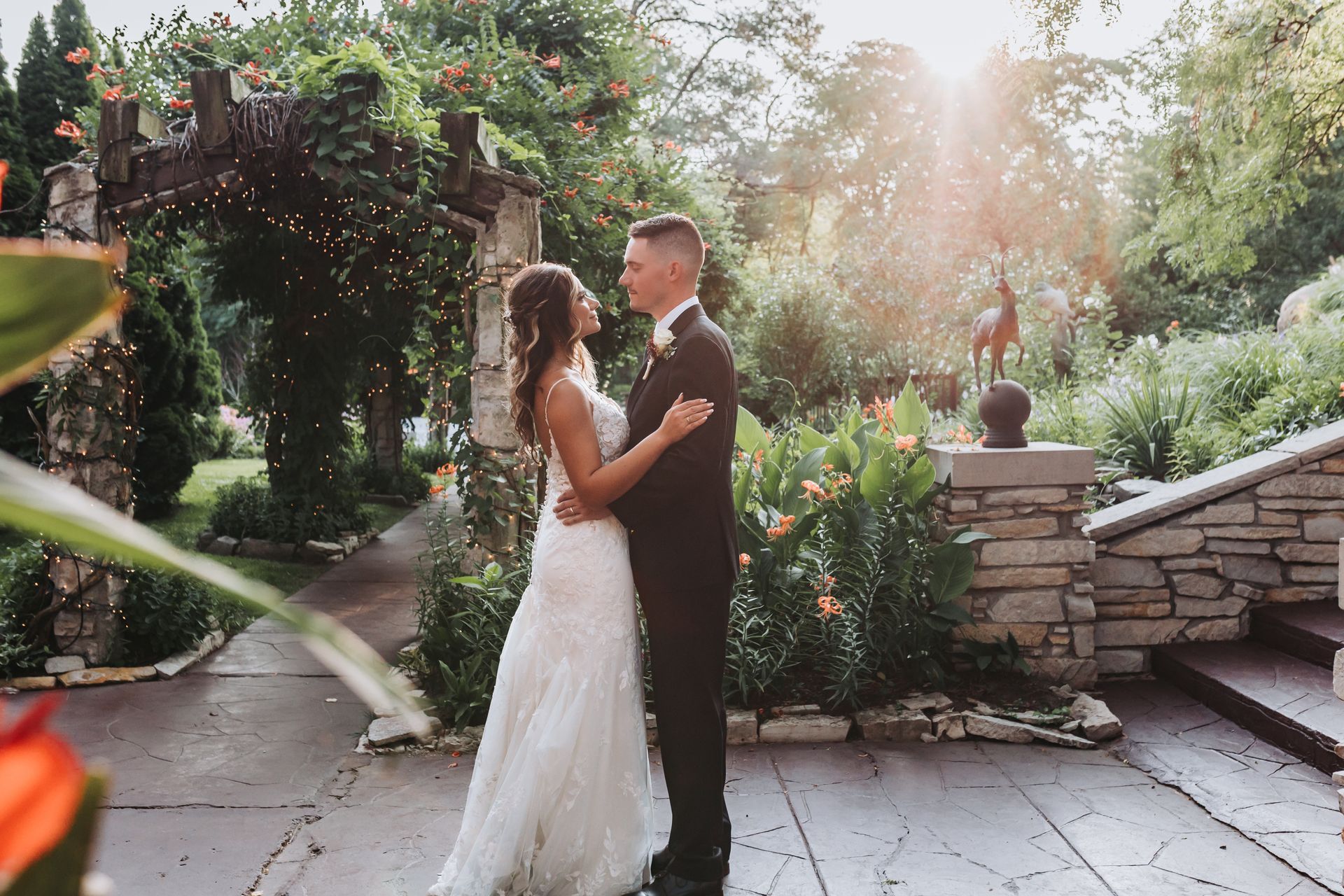 Couple embracing, wedding day, outdoors; sunlight flares, stone patio, greenery, romantic moment.