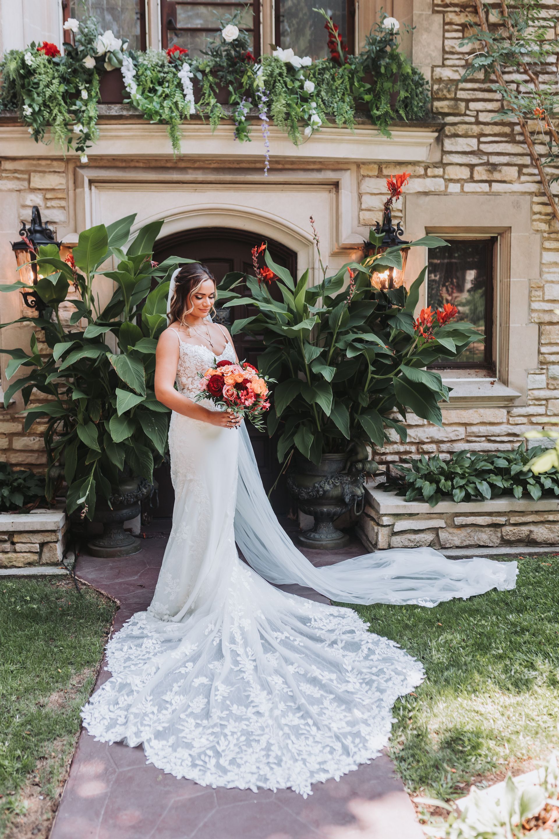 Bride in white lace wedding dress, holding bouquet, standing on a path in front of a stone building with green plants.