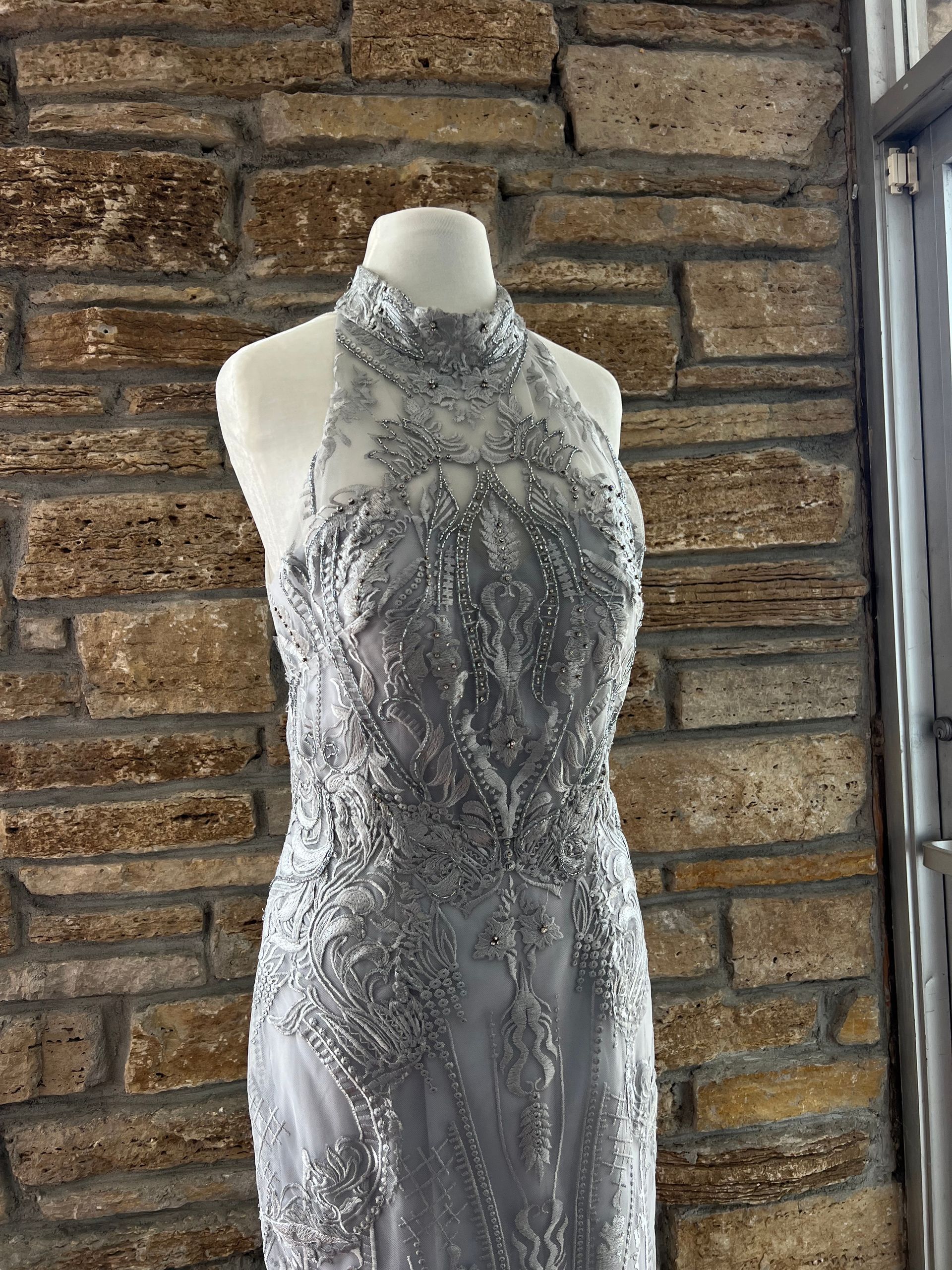 Silver formal gown on a mannequin, against a textured stone wall. High neckline, beaded details.