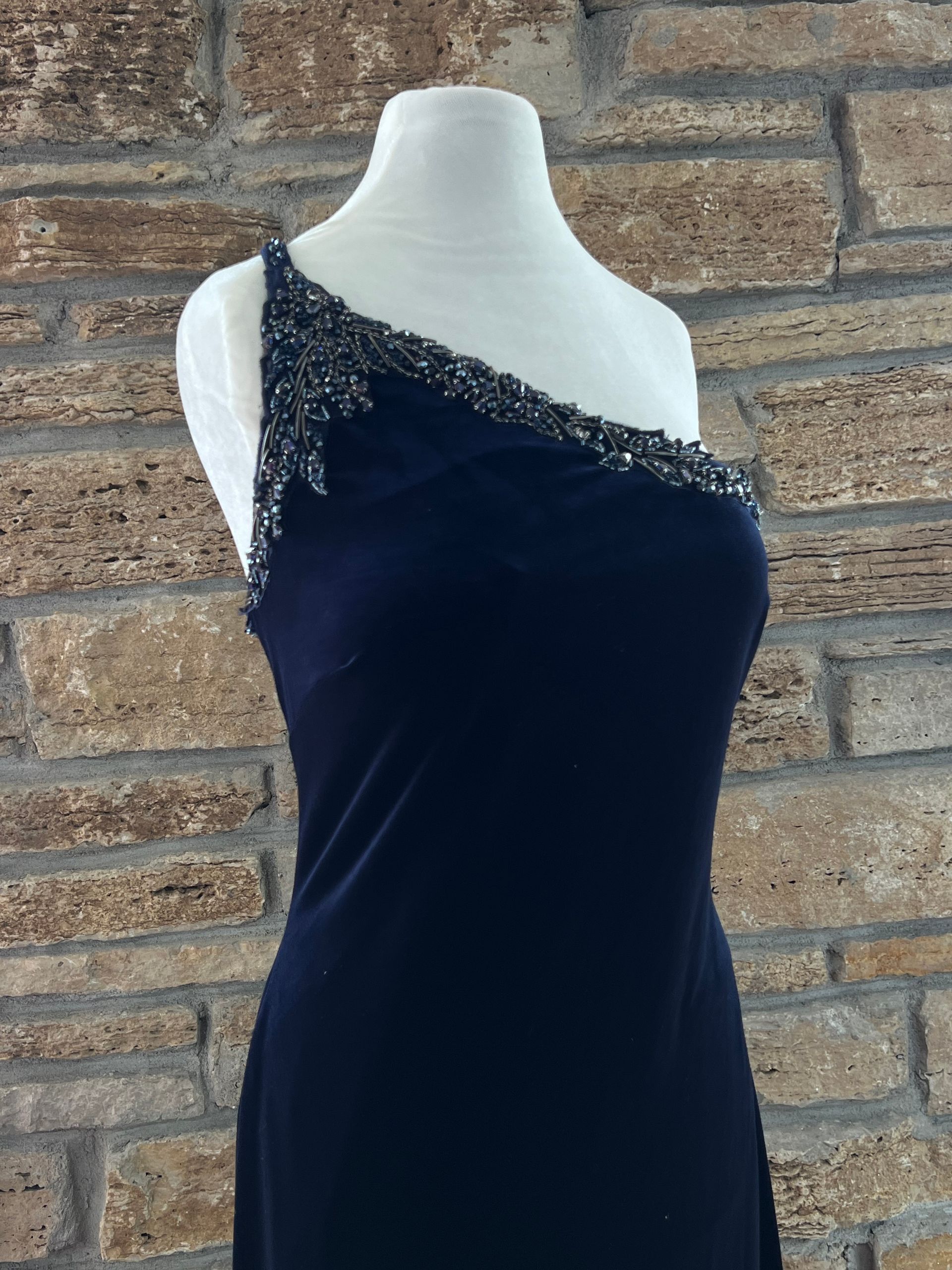 Navy blue one-shoulder formal dress with beaded embellishments displayed on a white mannequin against a brick wall.