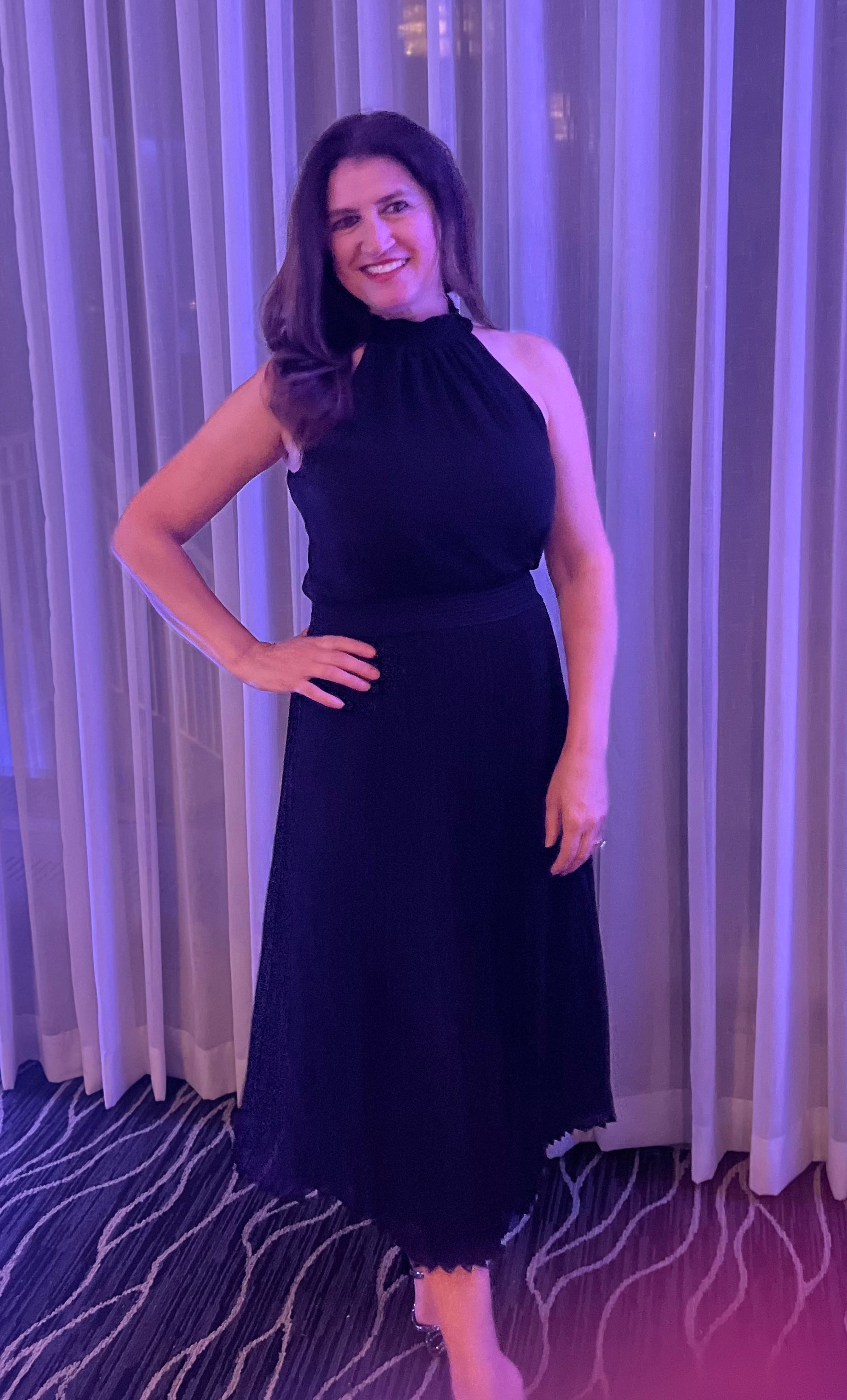 Fifi Papdopoulos in a navy dress is leaning against a curtain with her arm on her waist and smiling.