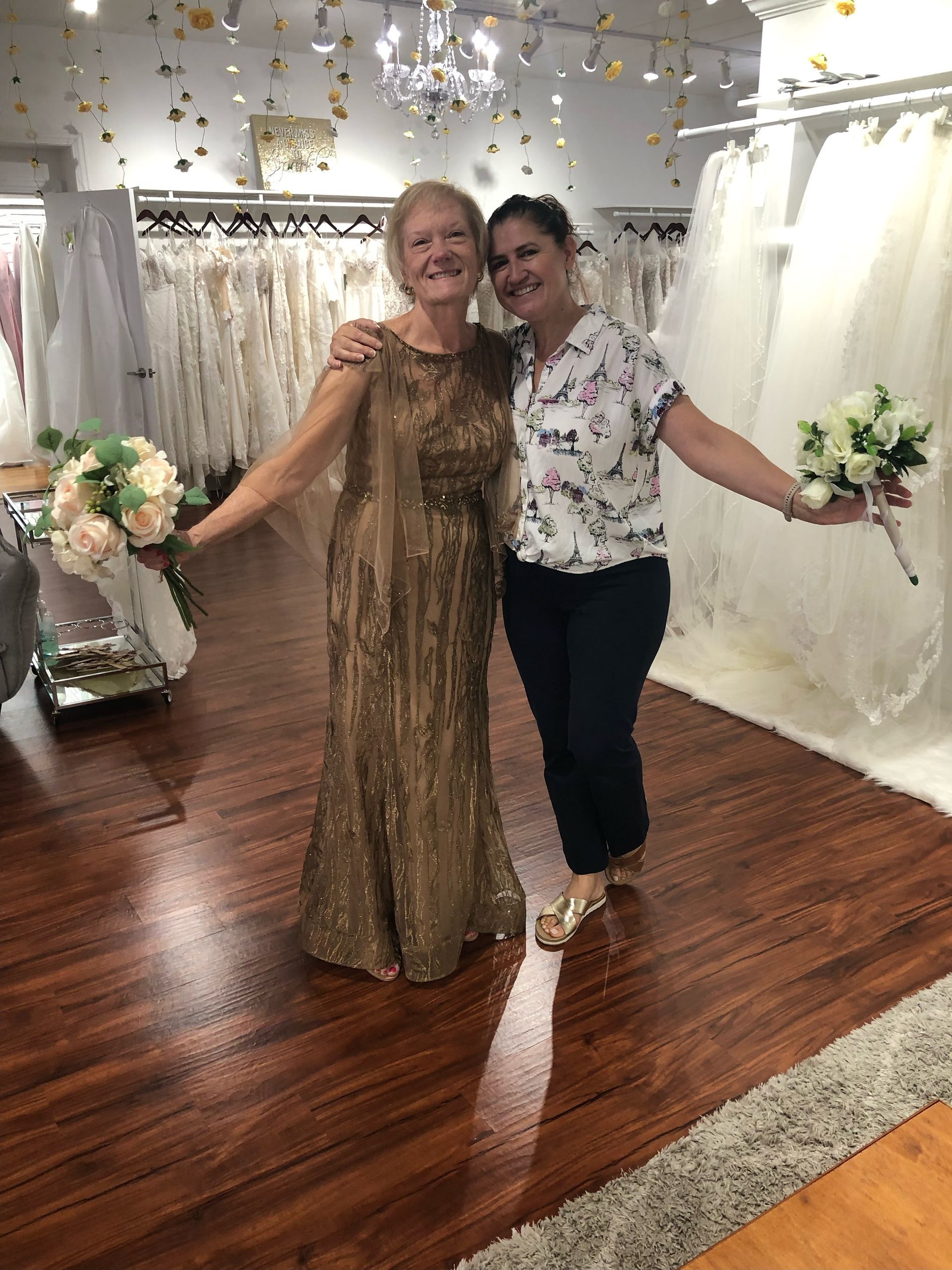 Two women in a bridal shop, holding bouquets, smiling. One in a gold gown, the other in a floral shirt.