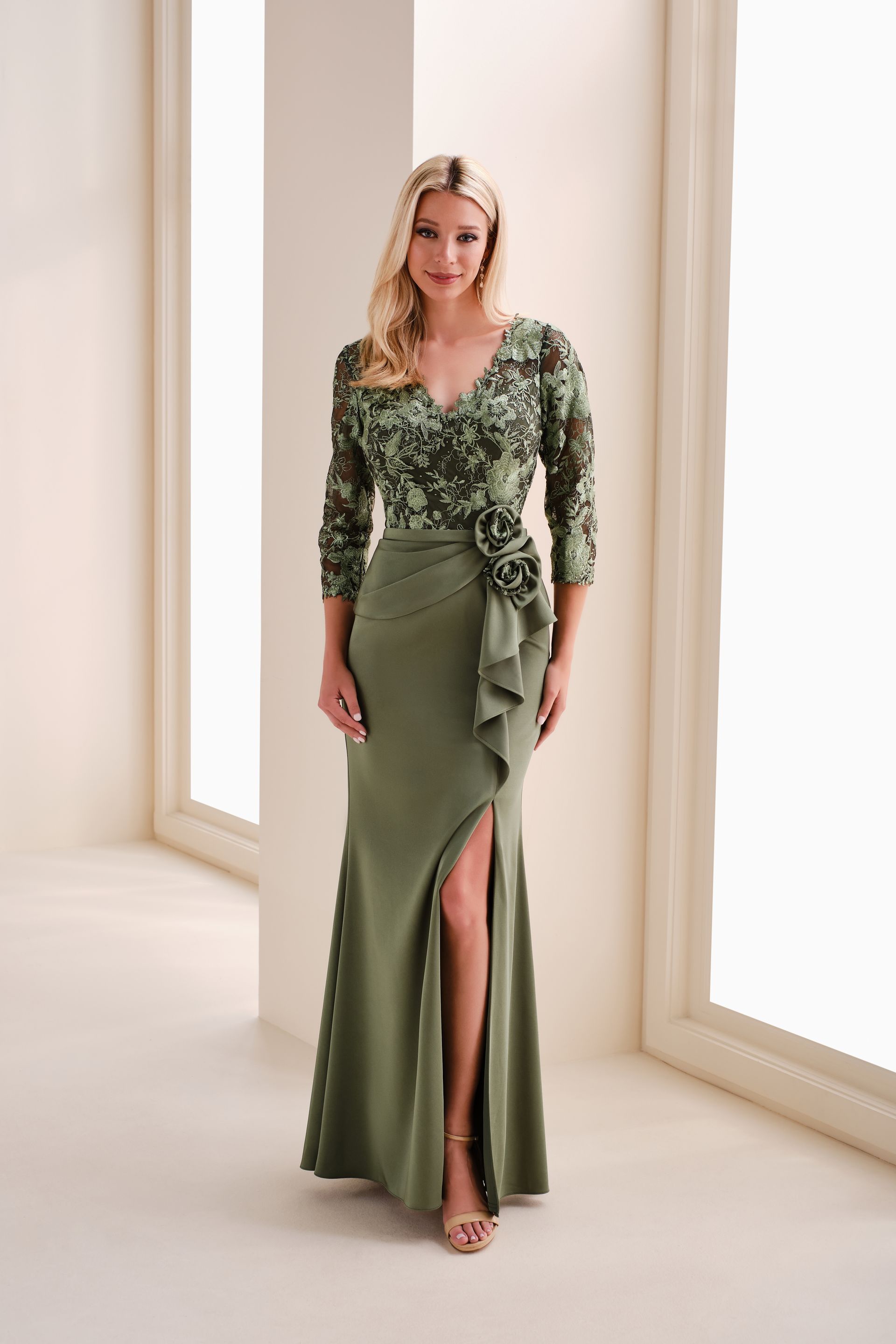 Woman in olive green formal dress with beaded top and thigh-high slit, standing by windows.