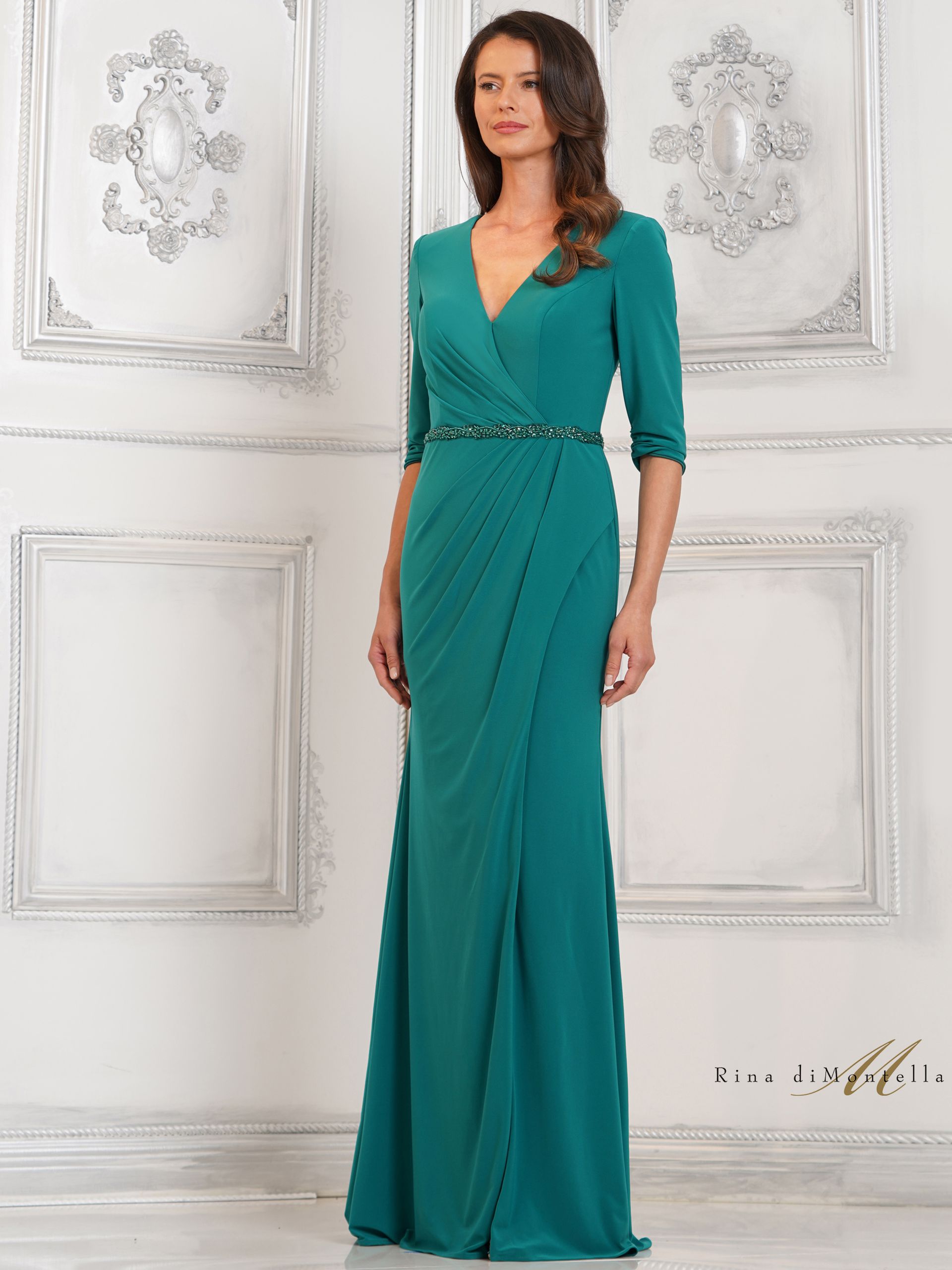 A woman in a long green dress is standing in front of a white wall.