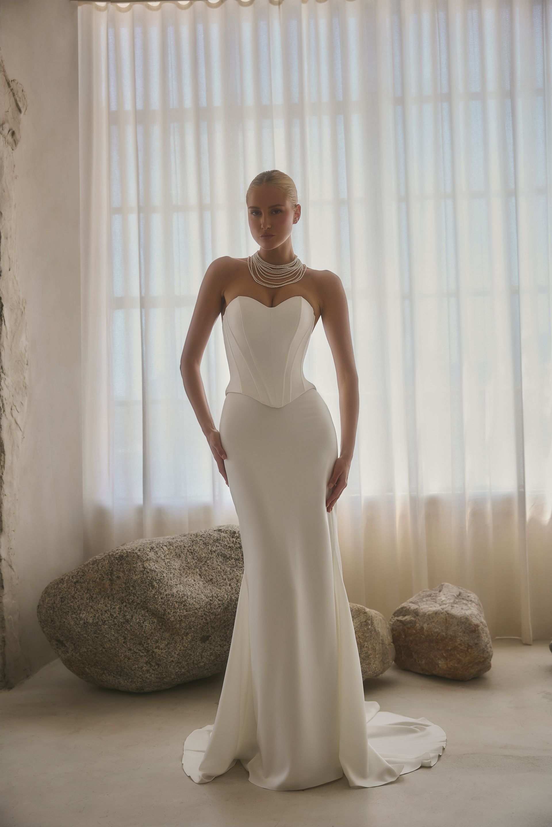 Woman in strapless white wedding dress, pearl necklace, posing in room with rocks, window.