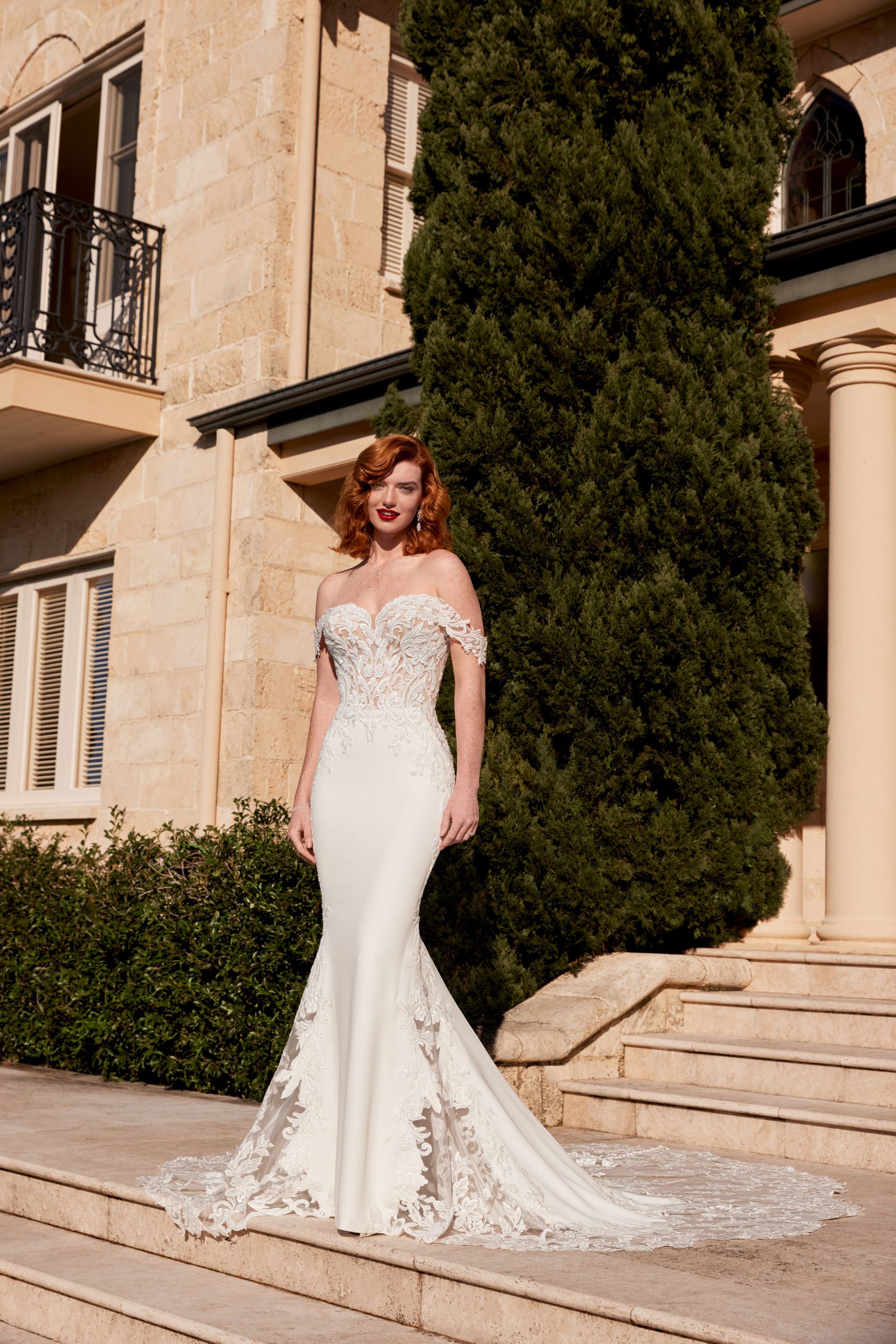 Woman in a white off-the-shoulder wedding dress stands on steps in front of a stone building with greenery.