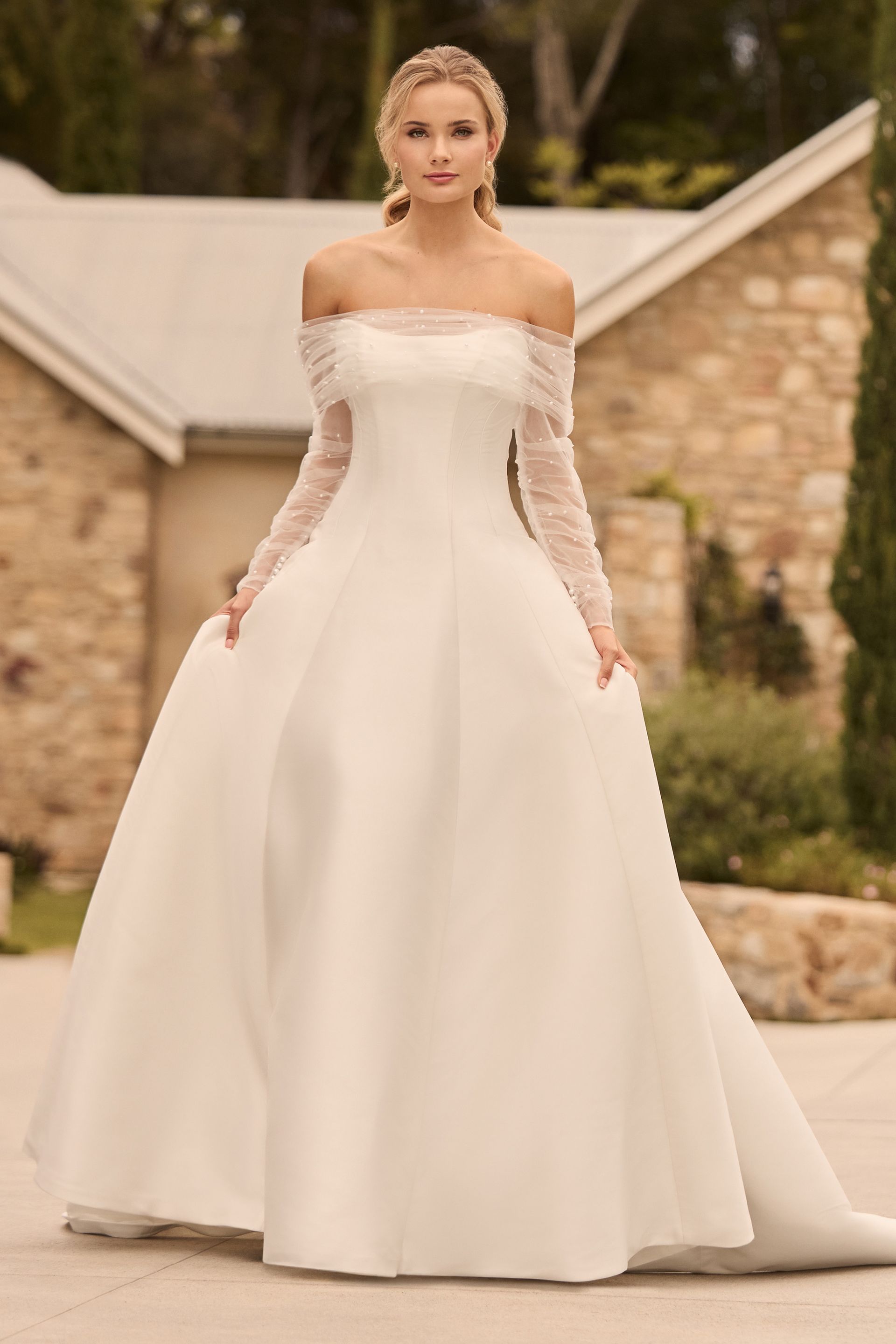 A woman in a white wedding dress is standing in front of a stone building.