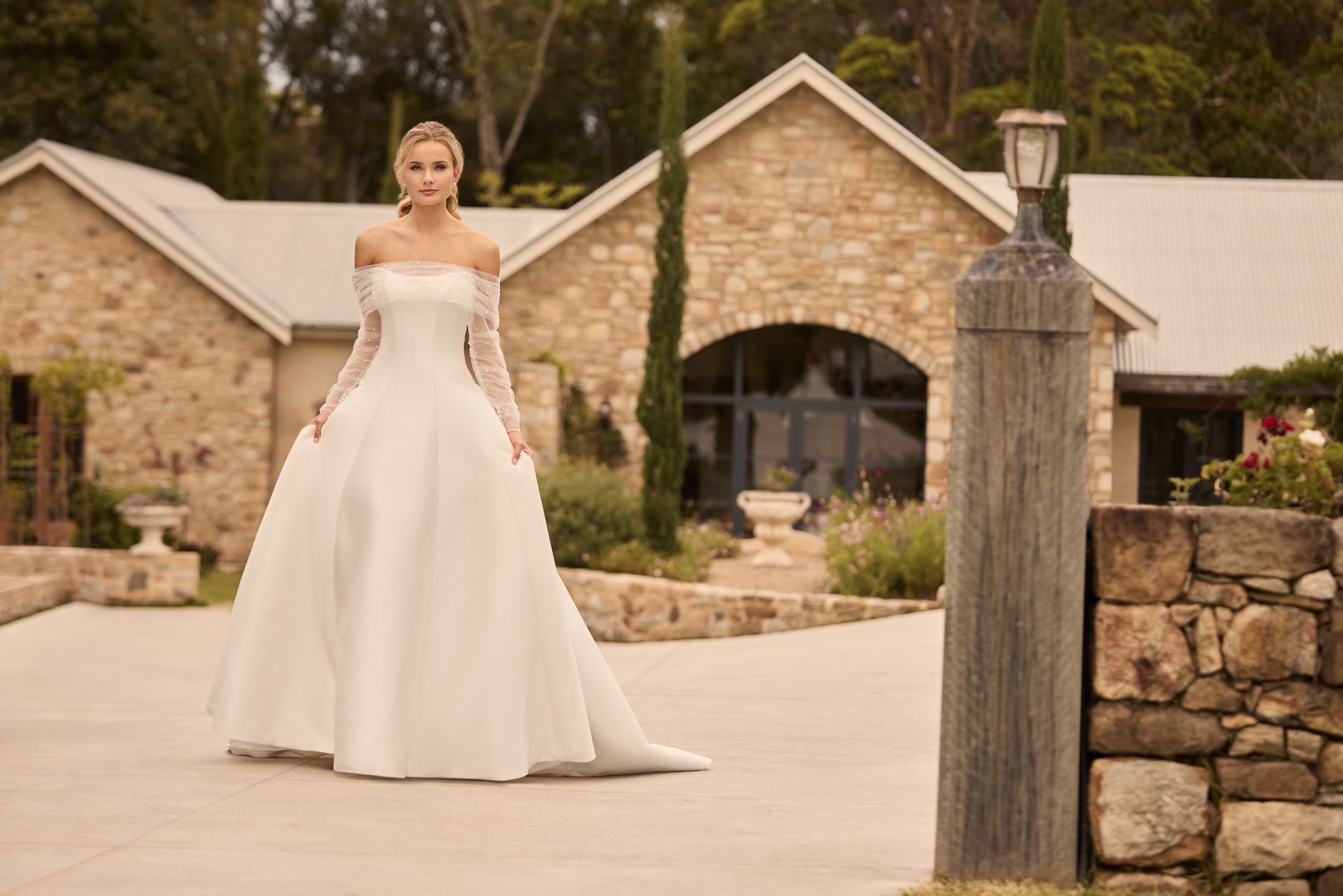 Woman in a white off-the-shoulder wedding dress stands outdoors in a garden setting. Woman in a white off-the-shoulder wedding dress stands outdoors in a garden setting.