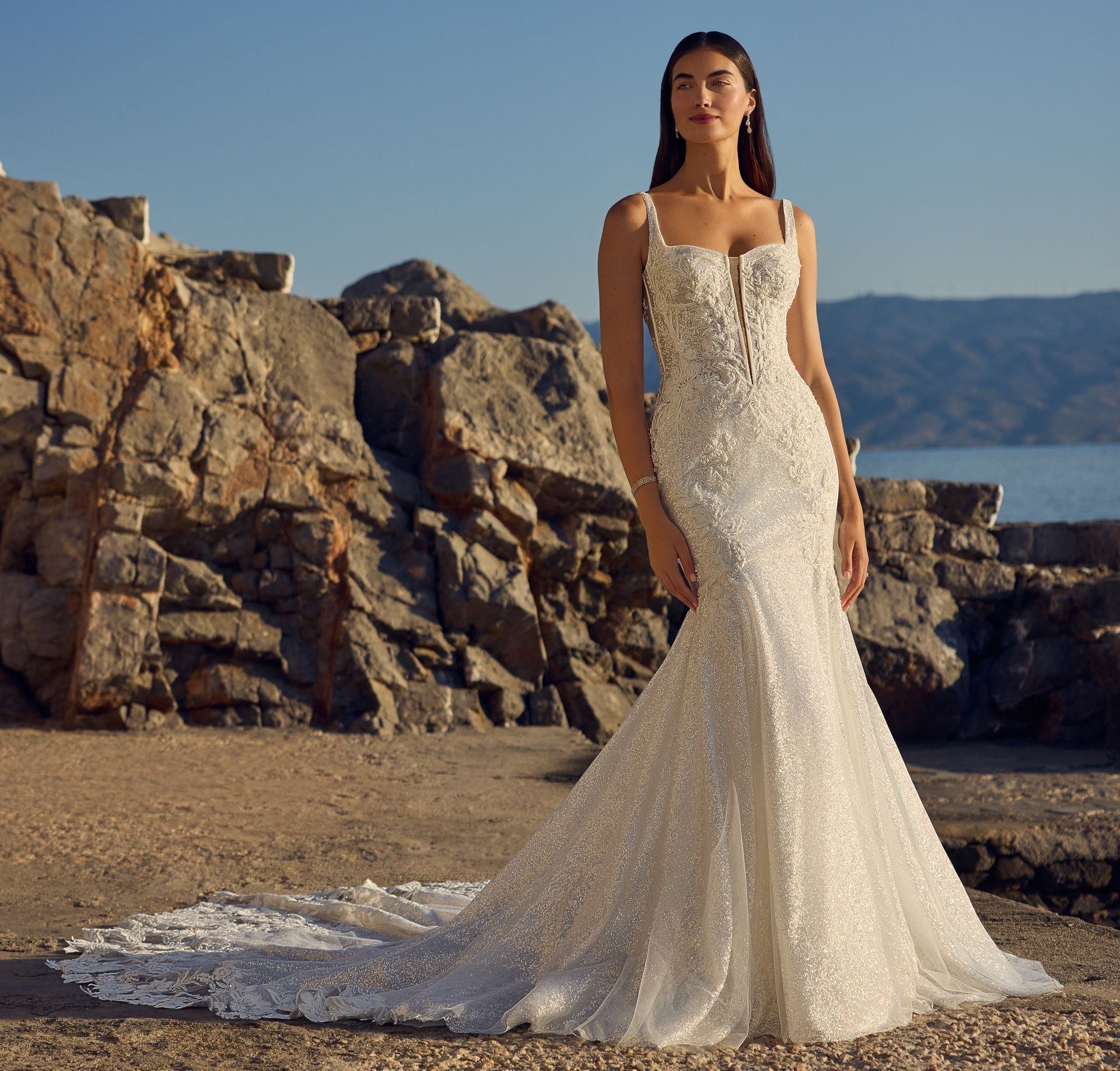 Woman in a sparkling white mermaid-style wedding dress with a long train, standing near rocks by the sea.