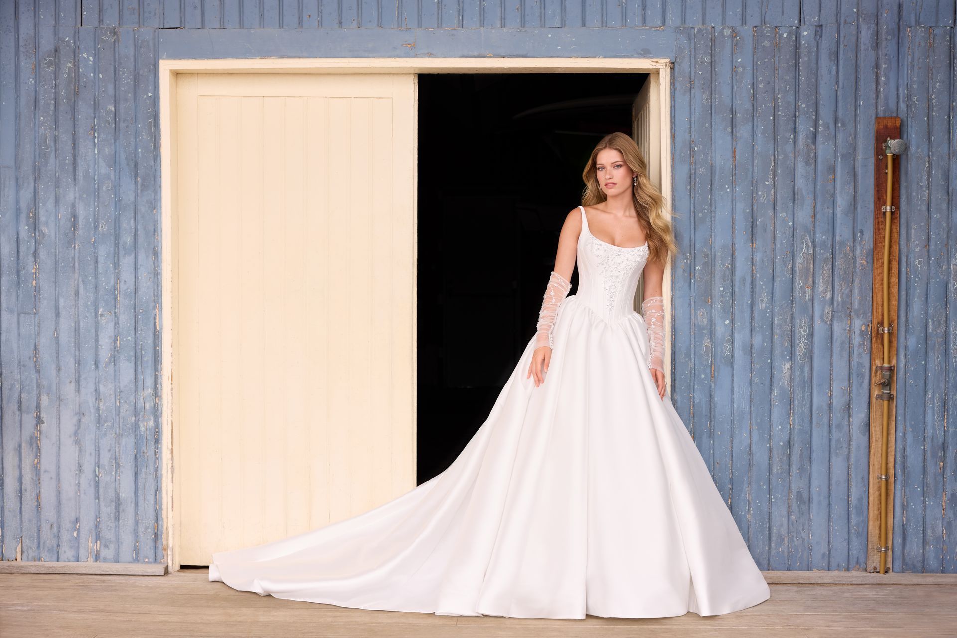 Woman in white wedding dress with long train stands in front of a barn door.