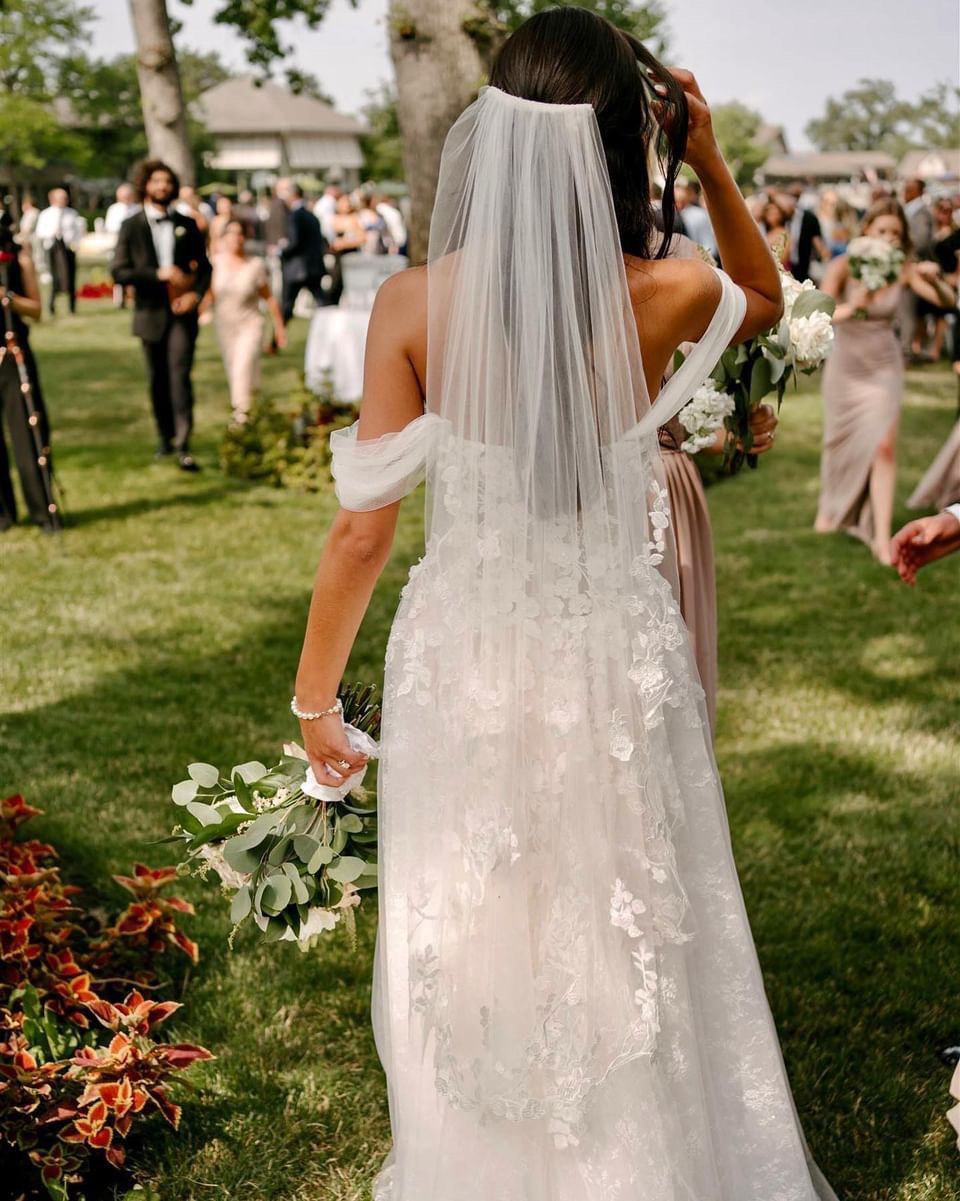 Bride in a white dress with veil, walking through a grassy wedding ceremony.