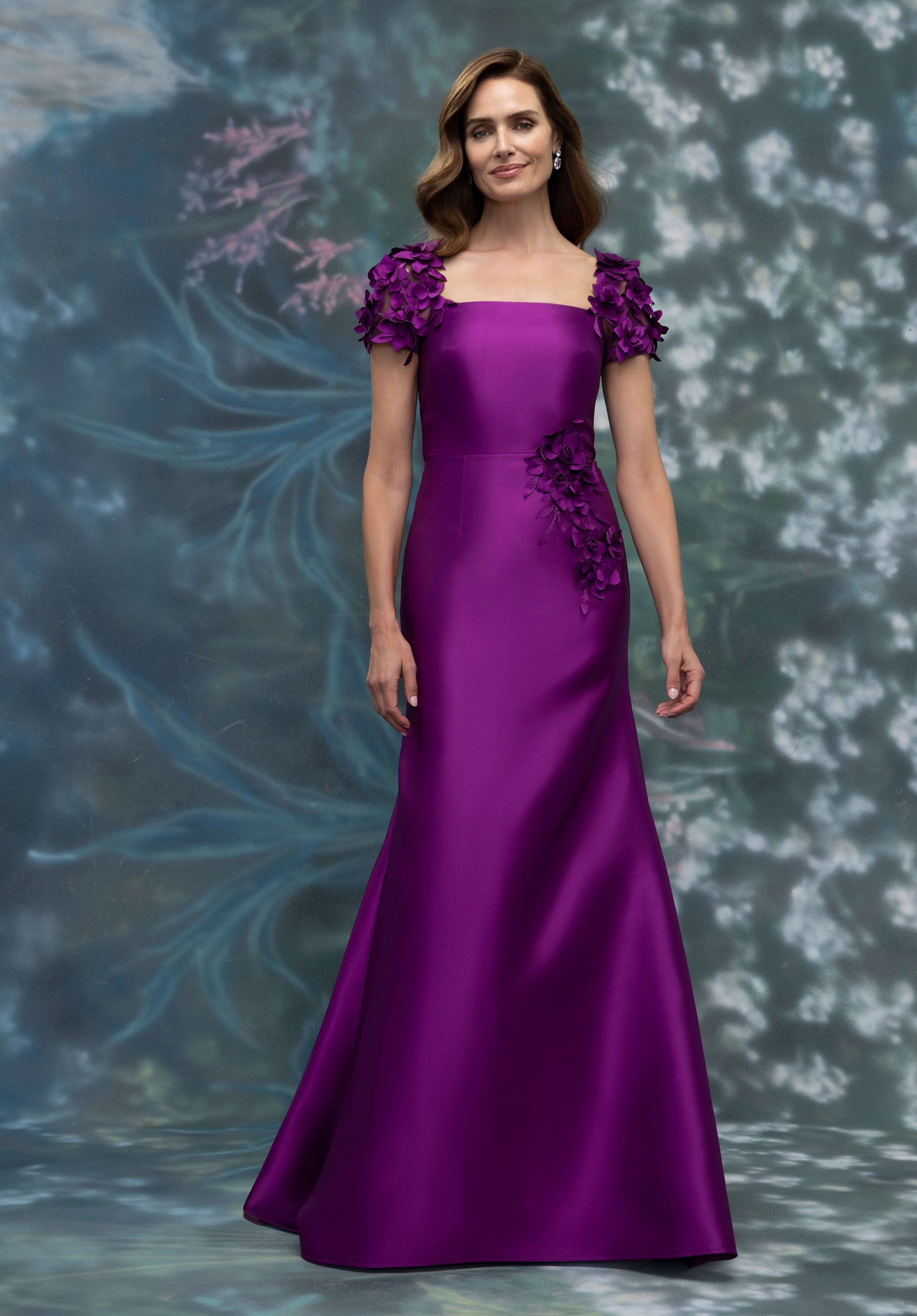Woman wearing a purple, floor-length dress with square neckline and floral shoulder details, standing in front of a blurred background.