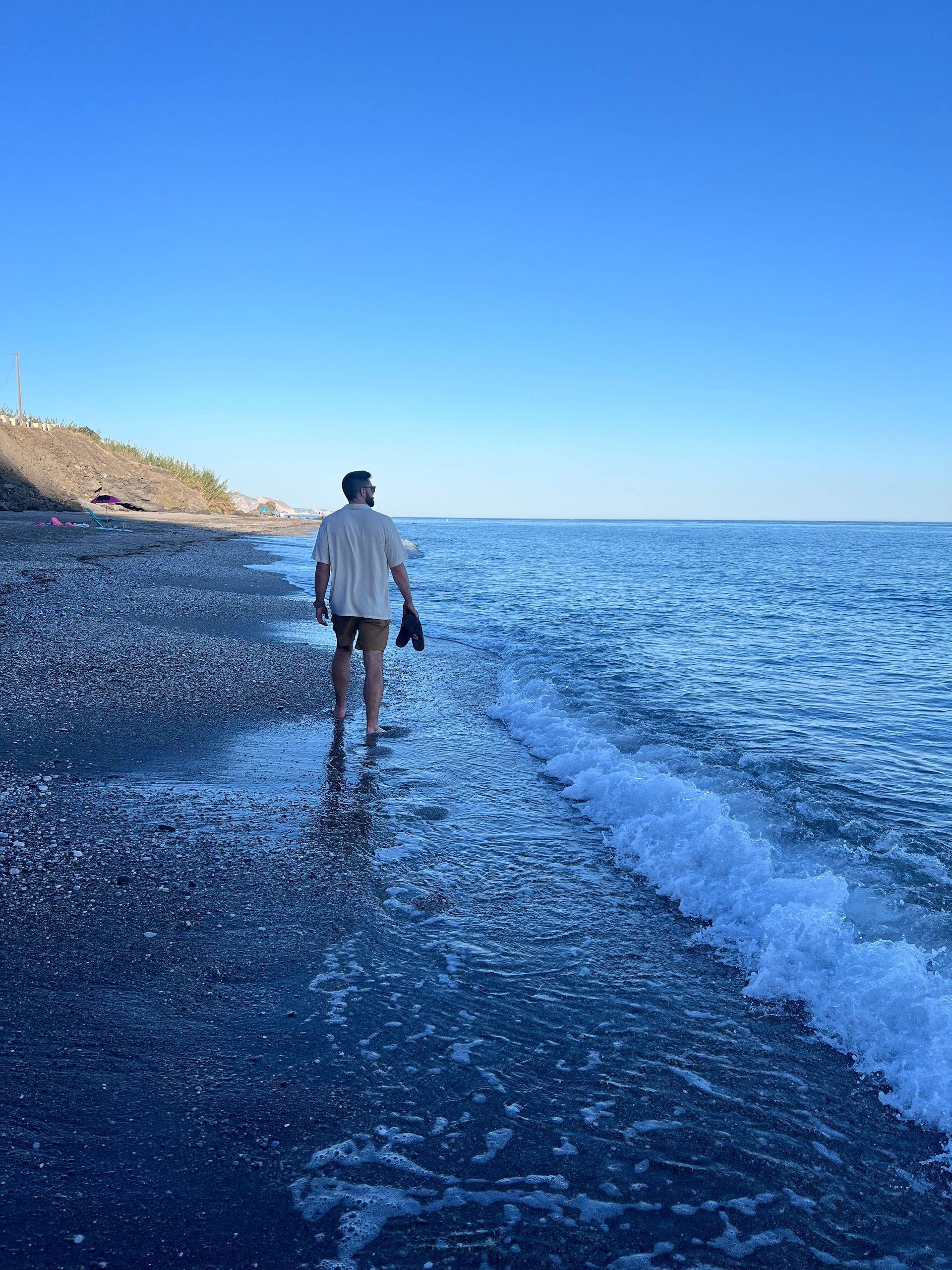 Sean walking on a dark pebble beach, looking out at the blue sea under a clear sky, holding shoes.