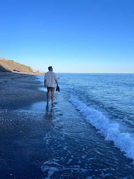 Sean walking on a dark pebble beach, looking out at the blue sea under a clear sky, holding shoes.