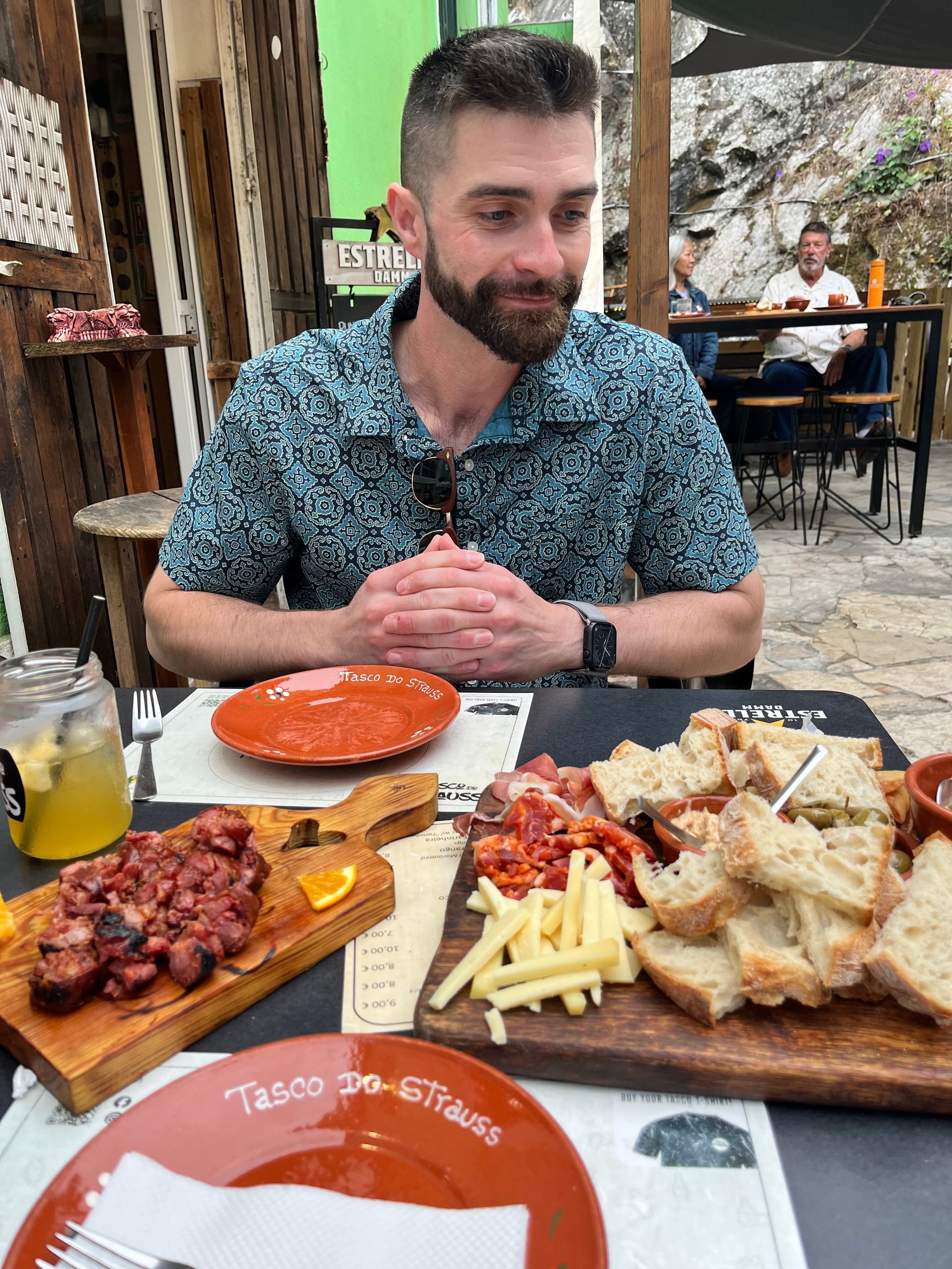 Sean seated at an outdoor table with food platters, looking down. Restaurant setting.