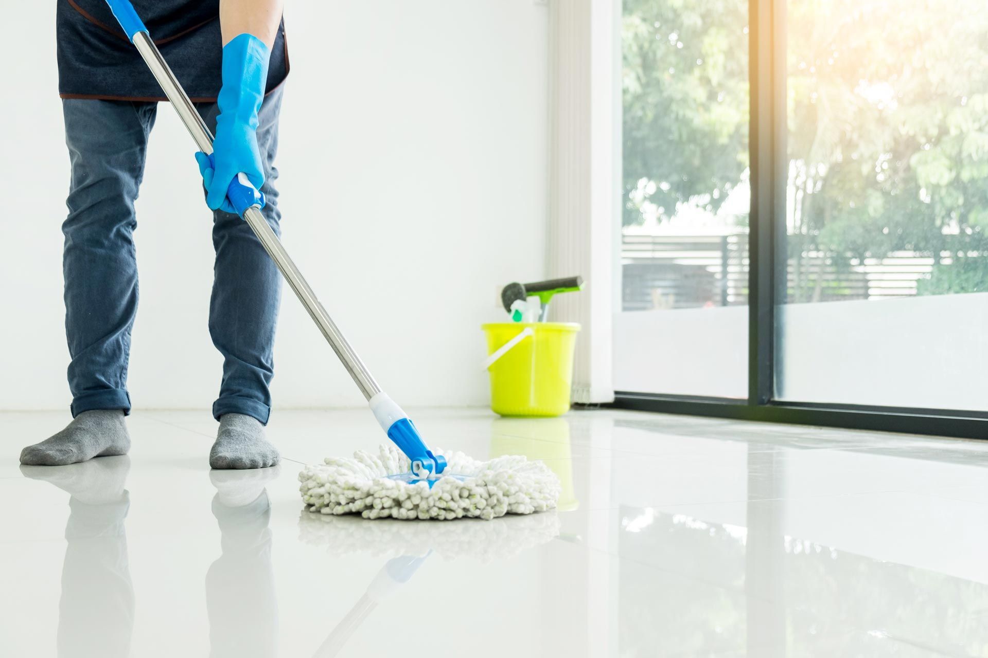 Person mopping a shiny white floor near a window, wearing blue gloves and jeans.
