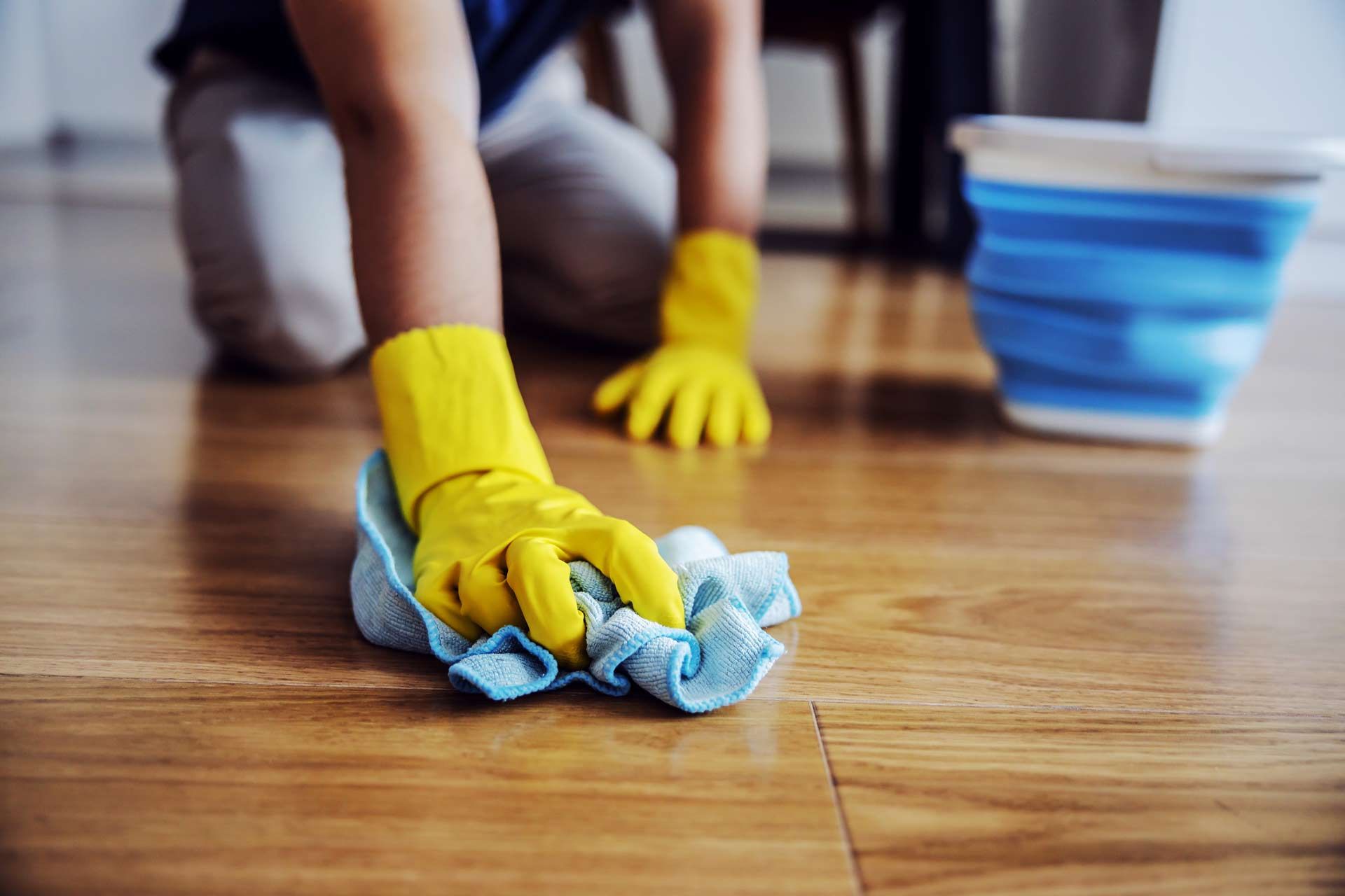 Person wearing yellow gloves wiping a wooden floor with a blue cloth; a blue bucket in the background.