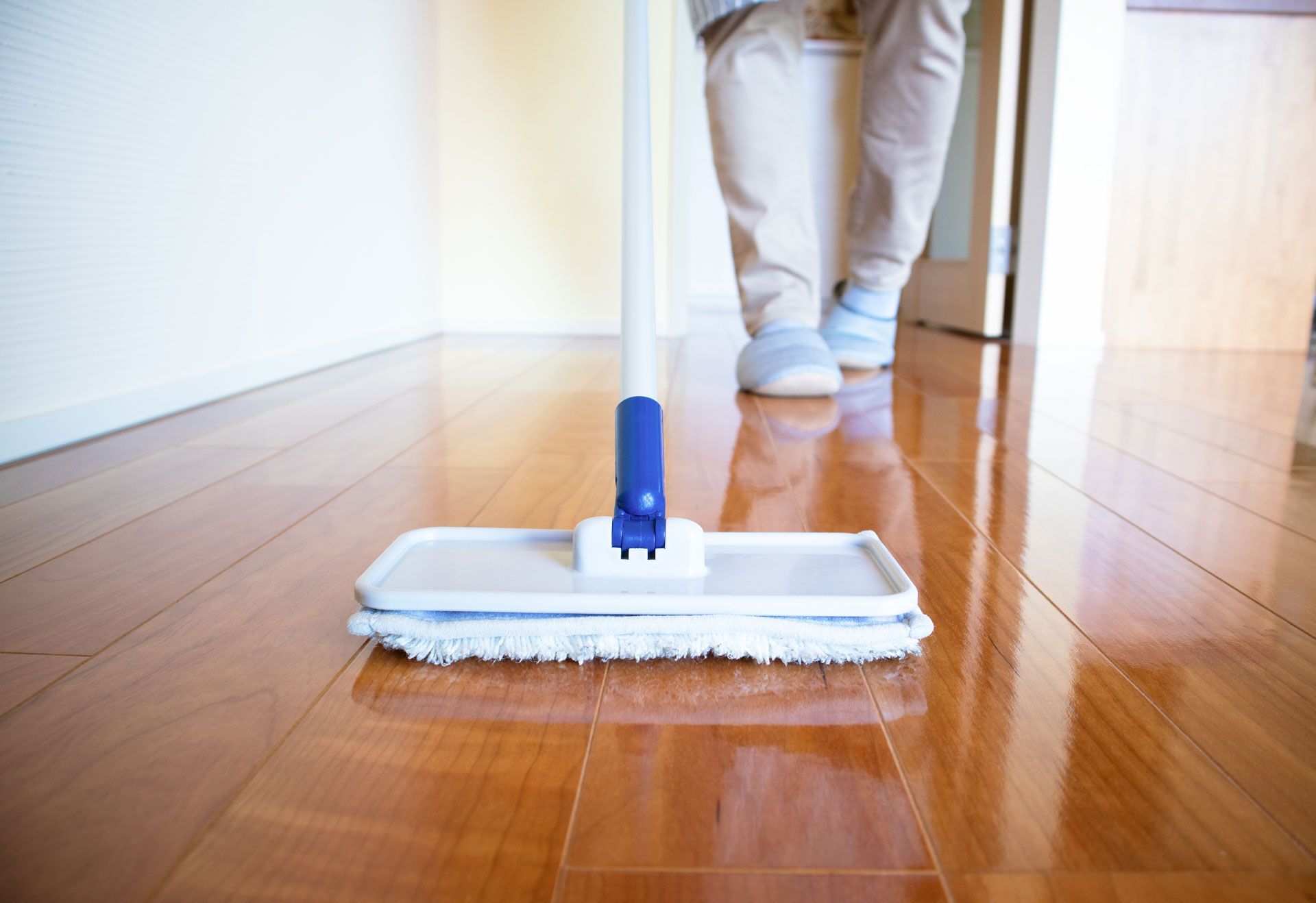 Person mopping a shiny, wooden floor with a white and blue mop; indoors.