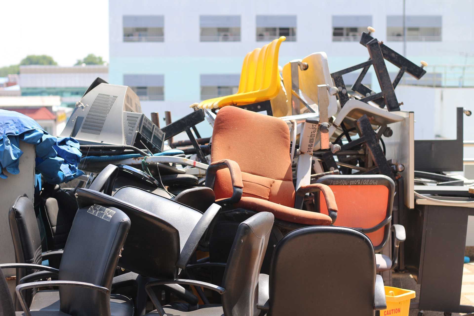 Pile of discarded office chairs and equipment in an outdoor setting; some chairs are orange, black, and yellow.