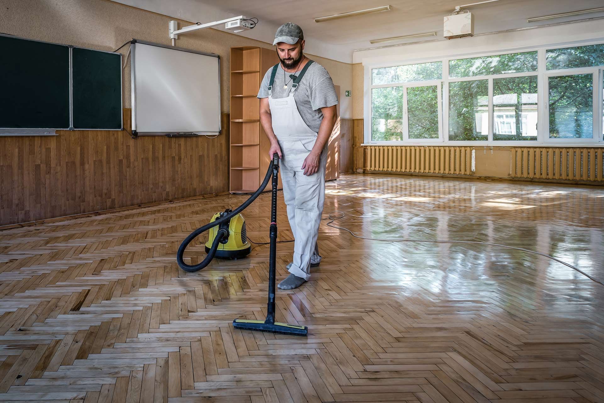 Person in overalls vacuums a wooden floor in a classroom with windows.